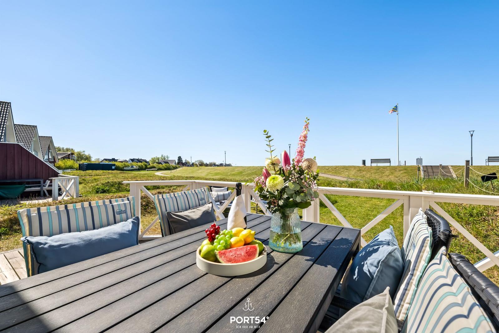 Terrasse mit Tisch, Stühlen und Obst. Blick auf grüne Wiese und Himmel.