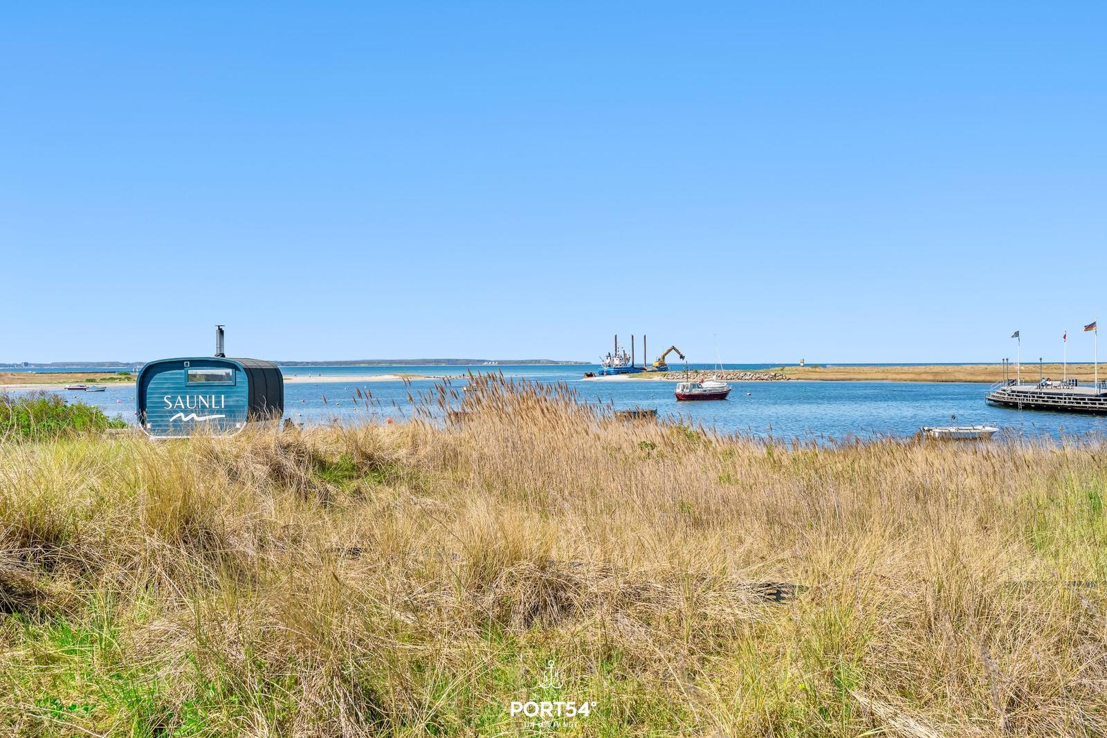 Blauer Saunli-Wohnwagen am Wasser mit Booten und Hafen im Hintergrund.