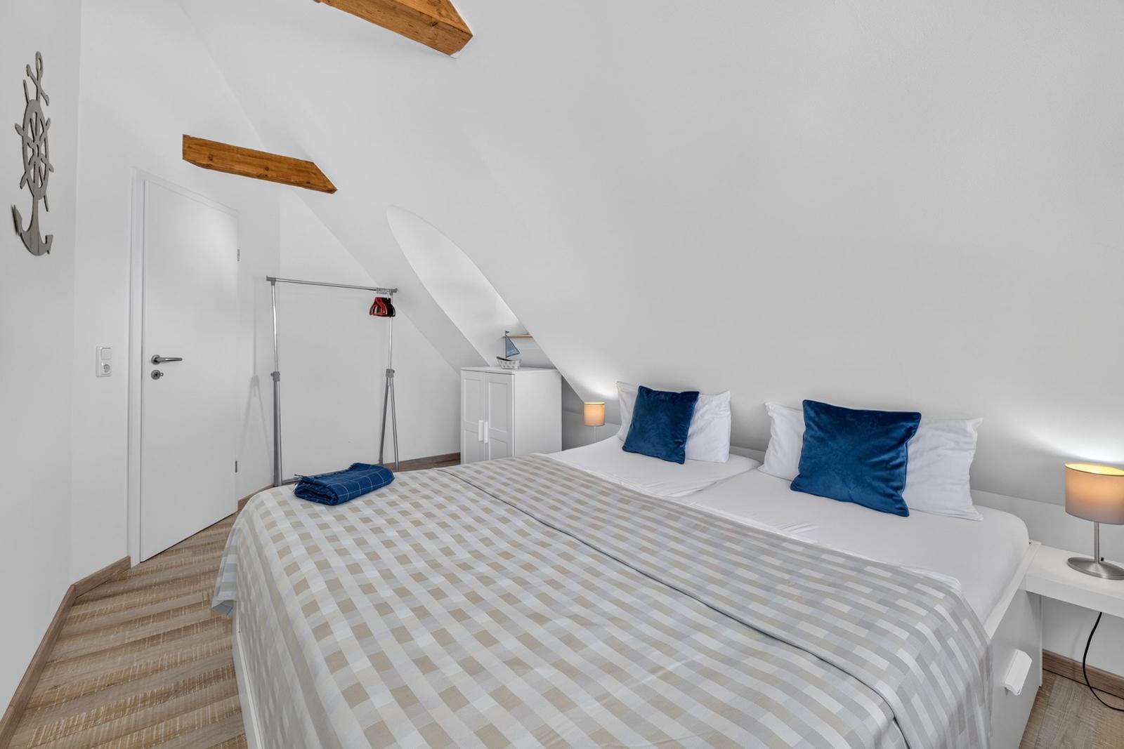 Schlafzimmer mit Dachschräge, Bett mit blauen Kissen und Holzbalken an der Decke.