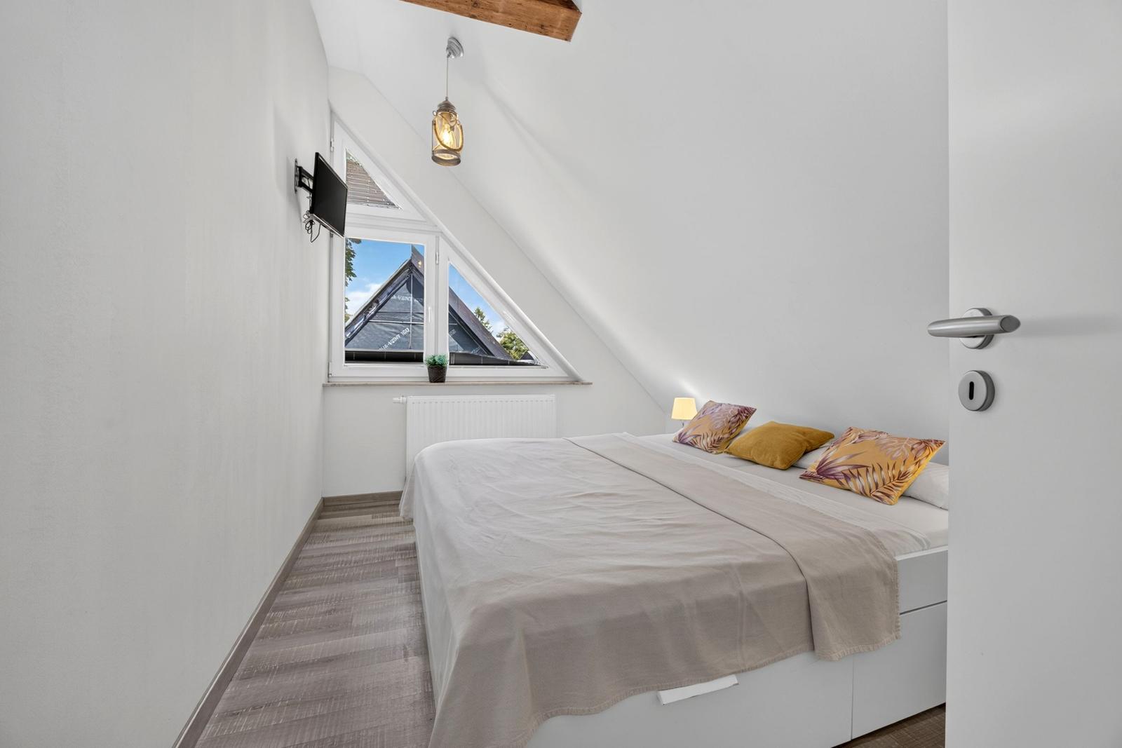 Schlafzimmer mit Dachschrägen, Bett, Fenster und Wand-TV.