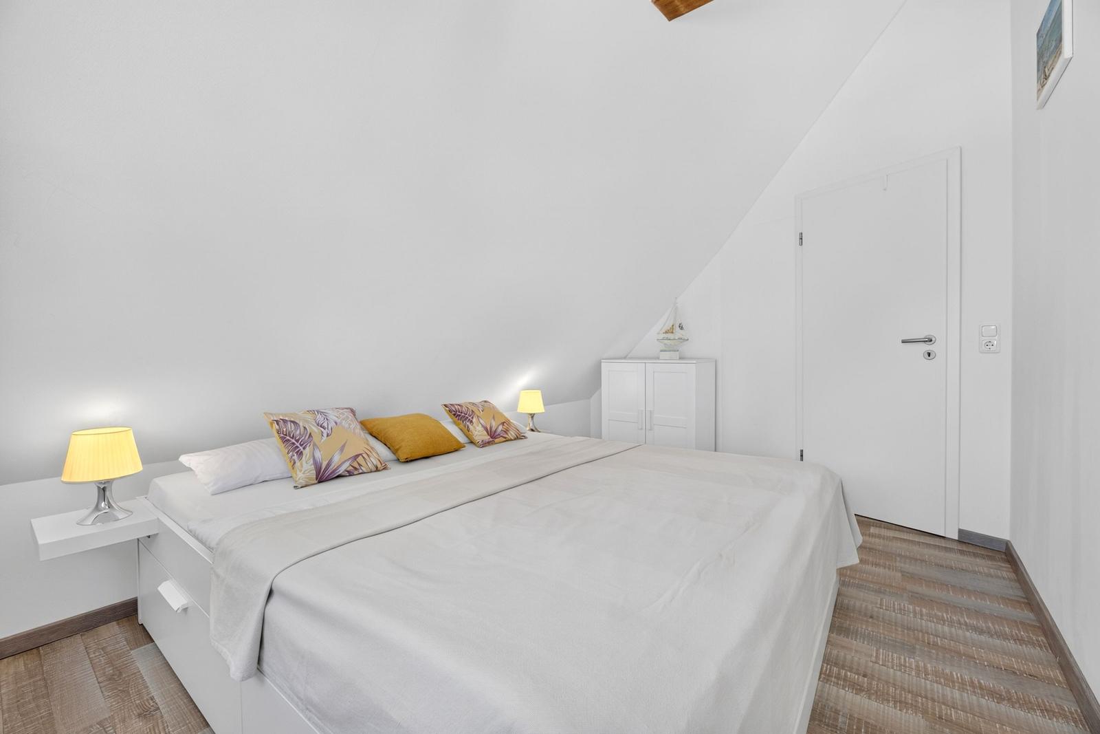 Schlafzimmer mit Dachschräge, weißem Bett und Holzboden. Zwei Nachttische mit Lampen.