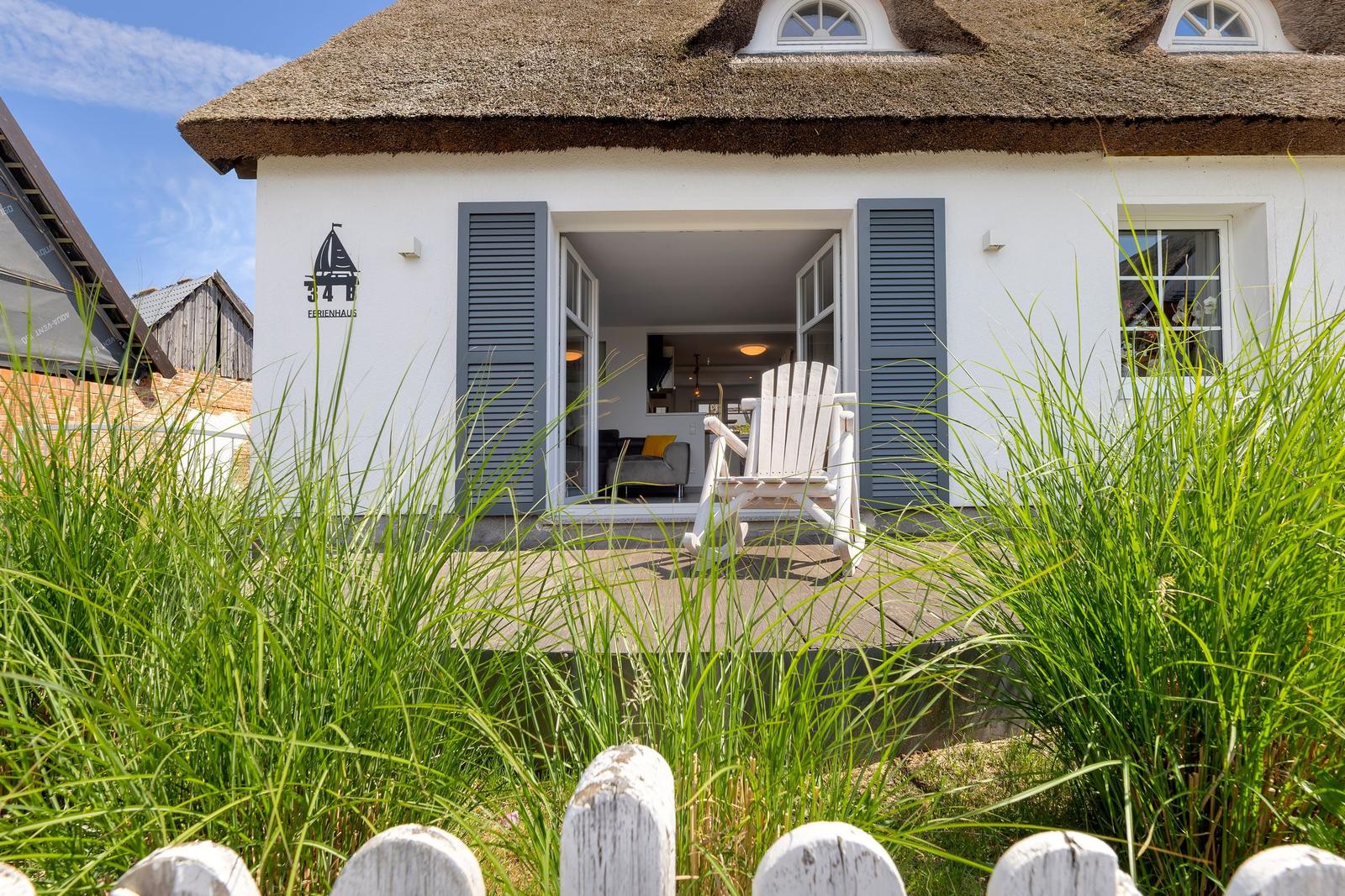 Ein weißes Ferienhaus mit Strohdach und offenem Eingang. Eine weiße Schaukelstuhl steht auf der Terrasse.