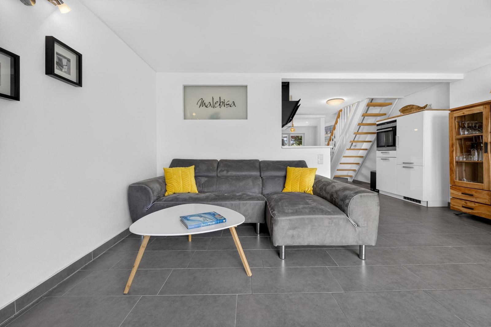 Wohnzimmer mit grauem Sofa, gelben Kissen und weißem Couchtisch. Küche und Treppe im Hintergrund.