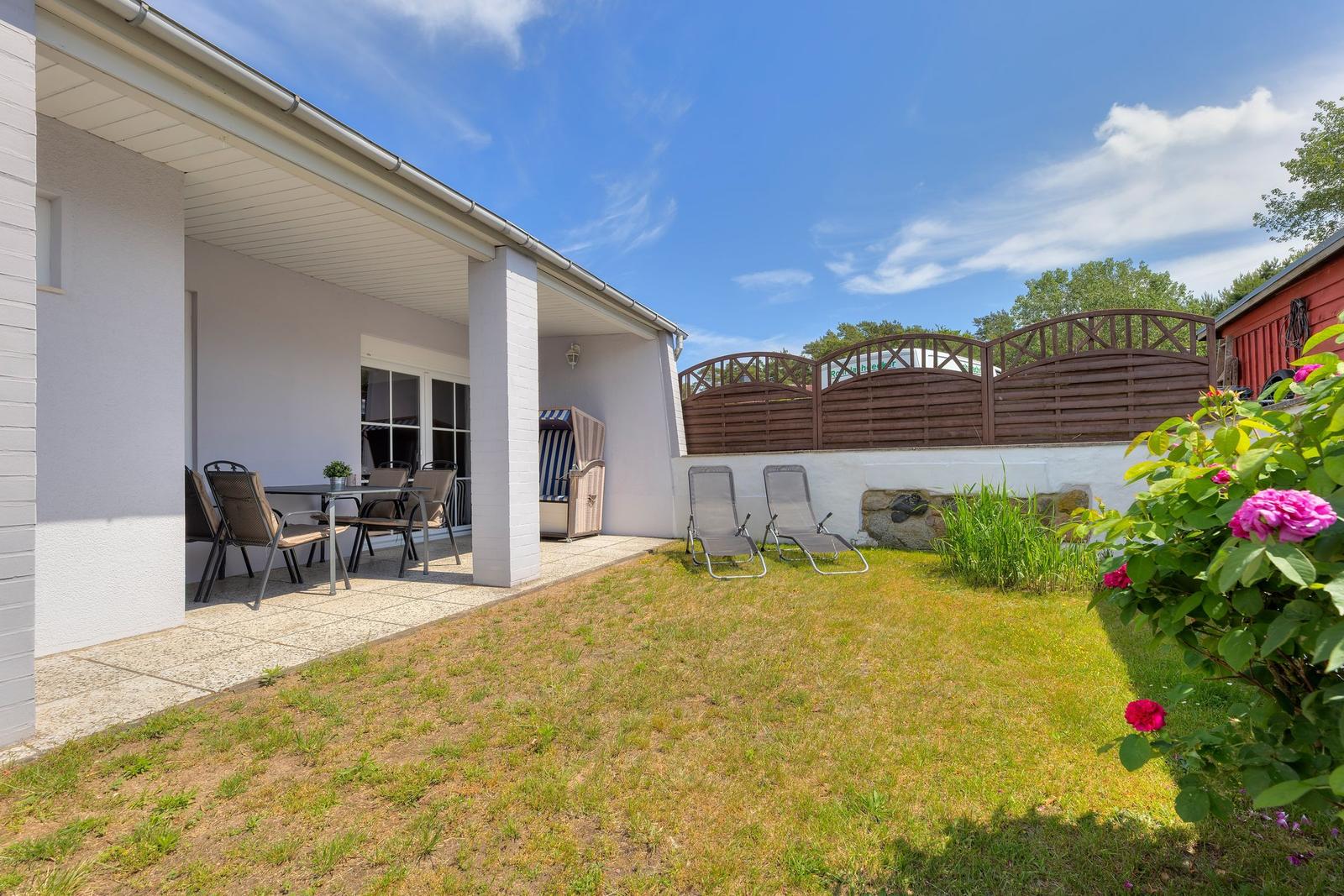 Terrasse mit Tisch und Stühlen, zwei Liegestühle auf dem Rasen, Blumensträucher und Holzgeländer.