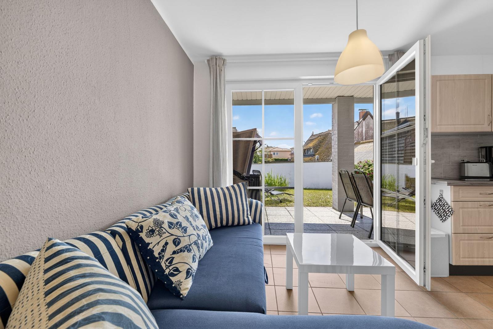 Ein Wohnzimmer mit Sofa, Tisch und Terrasse. Durchgehende Glastüren führen auf die Terrasse.