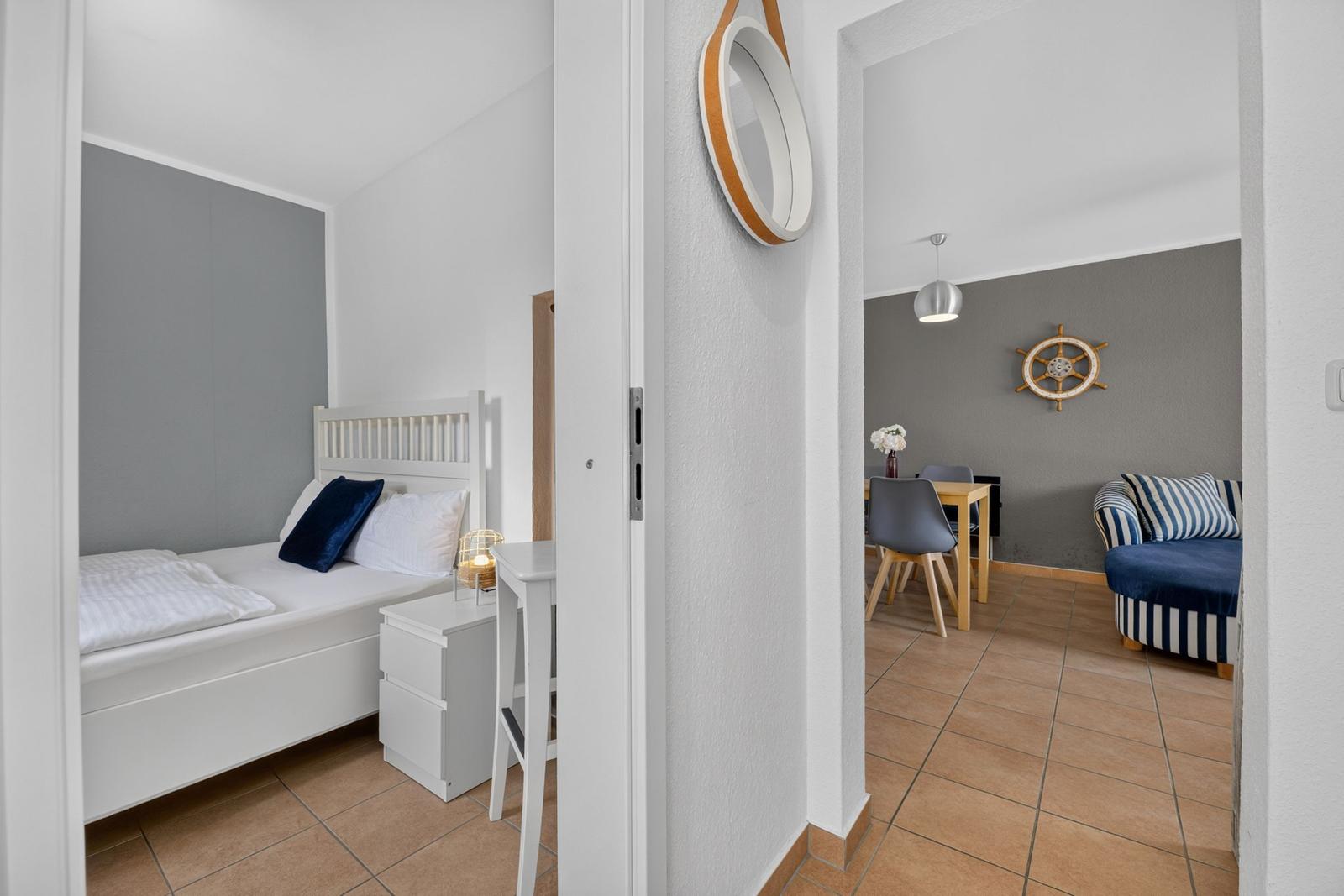 Schlafzimmer mit weißem Bett und Nachttisch. Durchgang in Wohnbereich mit Tisch, Stühlen und Sofa.