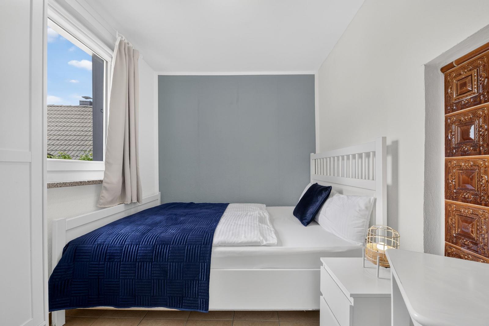Schlafzimmer mit weißem Bett, blauer Decke und Fenster mit Blick auf Dach.