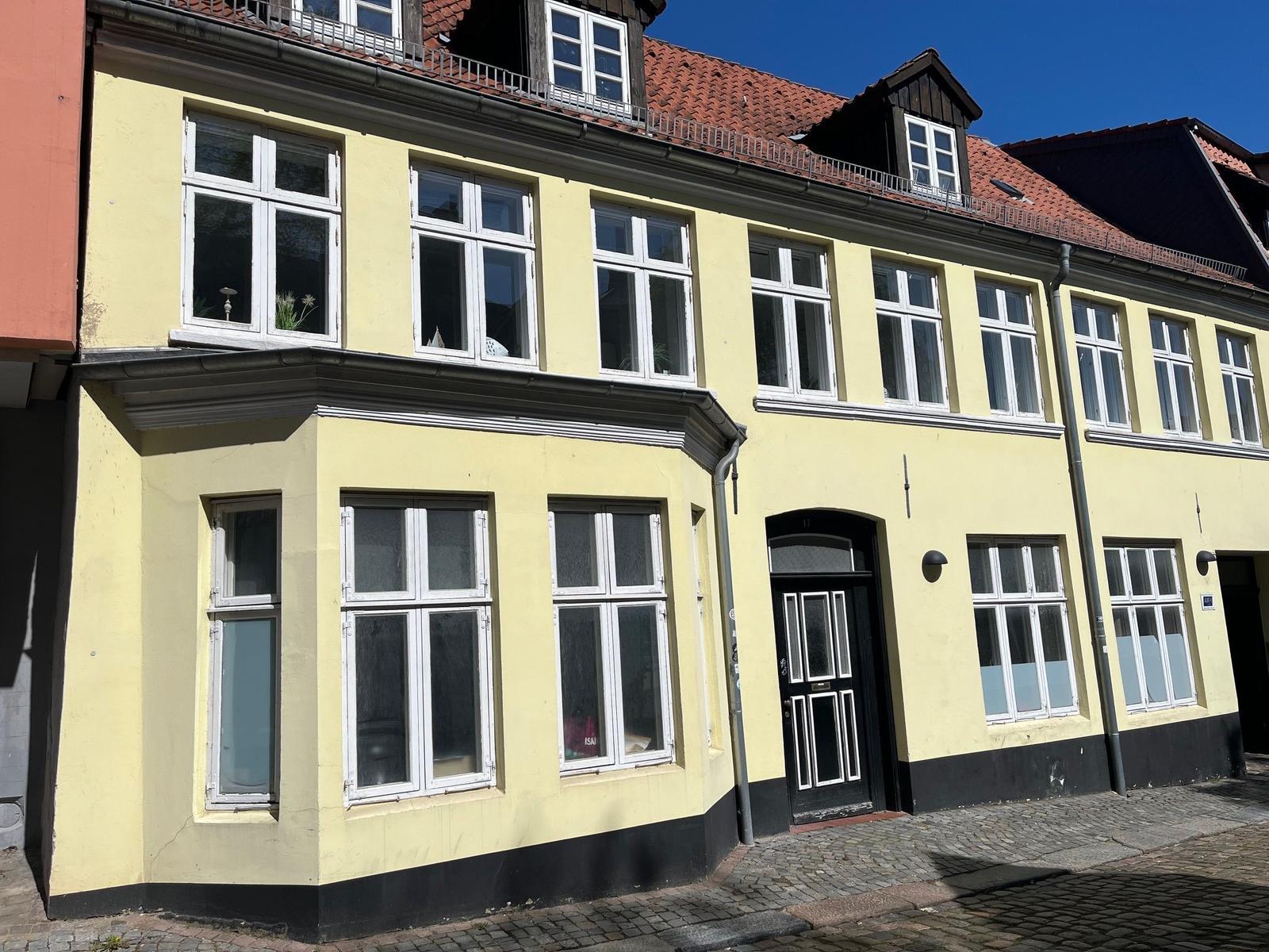 Ein gelbes Gebäude mit weißen Fenstern und rotem Dach. Eingangstür und Klinkerstraße sind sichtbar.