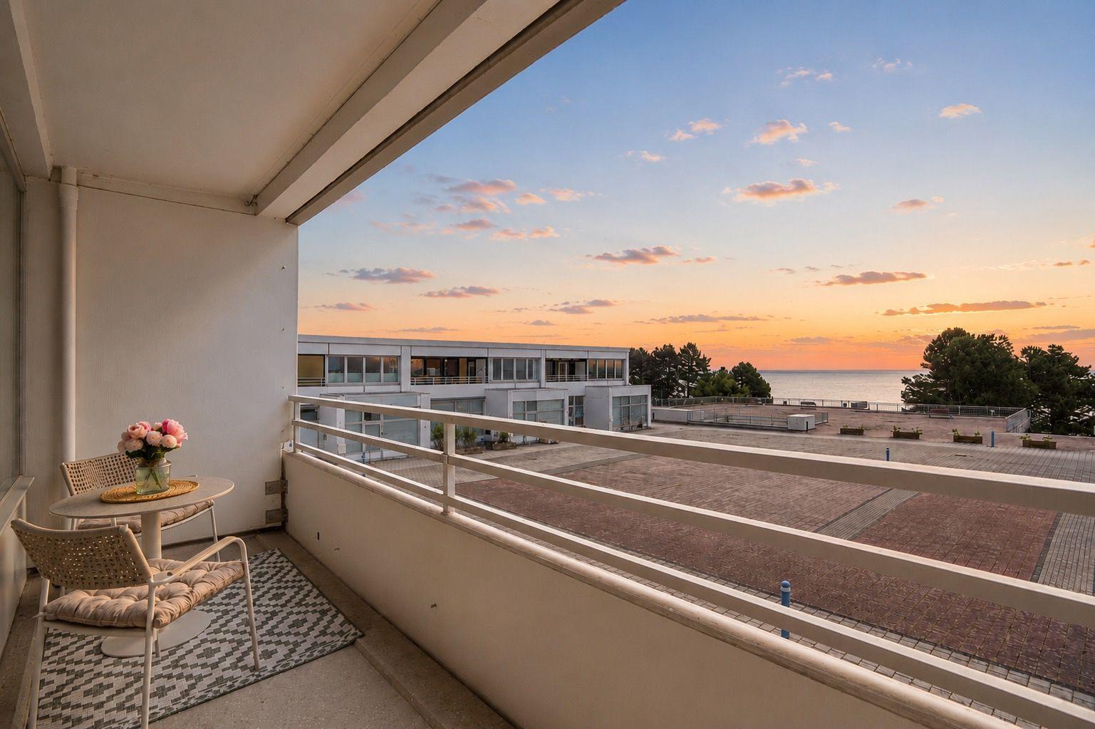 Balcony with table and chairs overlooking sea at sunset.