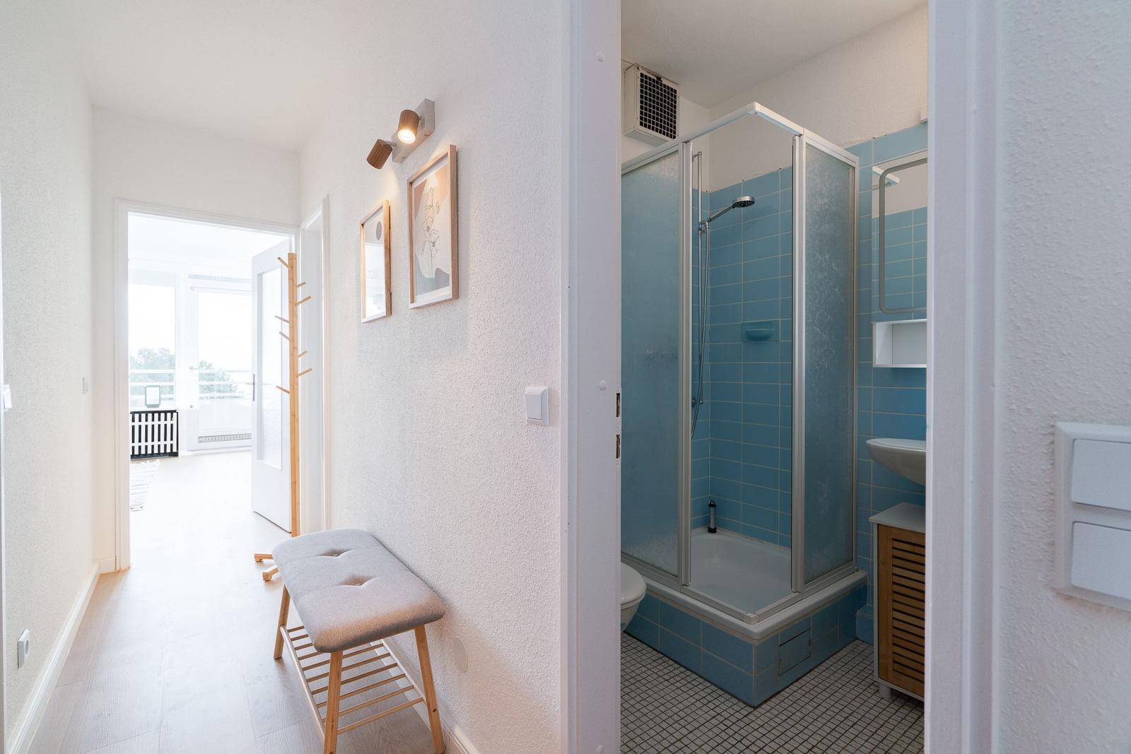 Hallway with stool and pictures, bathroom with shower and blue tiles.