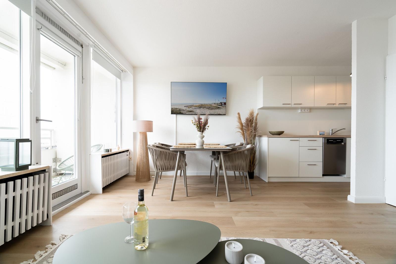 Dining area with table and chairs. Kitchen with cabinets and dishwasher. Large windows and wooden floor.