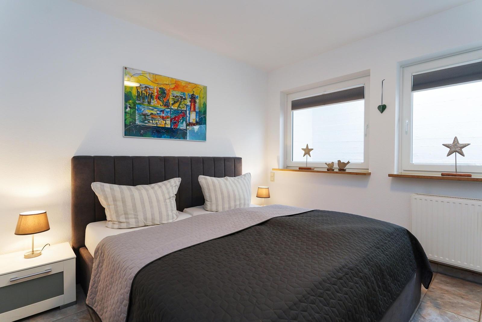 Schlafzimmer mit Bett, Nachttisch und Fenstern. Wandbild und Dekorationen sind sichtbar.
