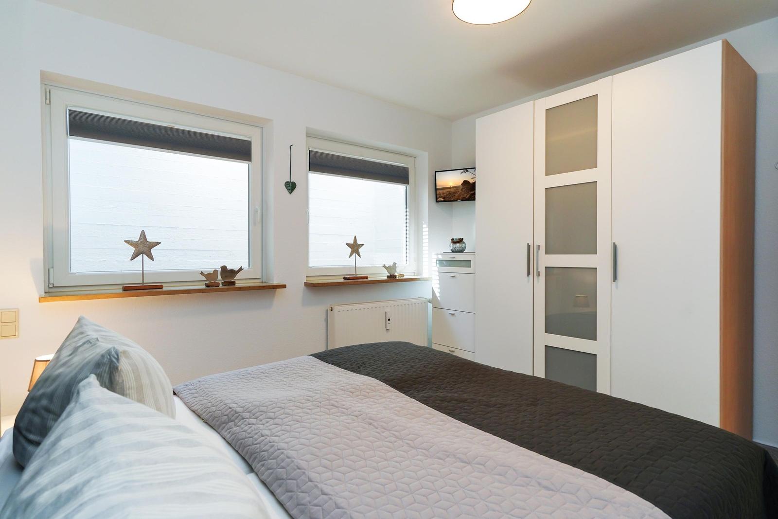 Schlafzimmer mit Doppelbett, zwei Fenstern und weißen Schränken.