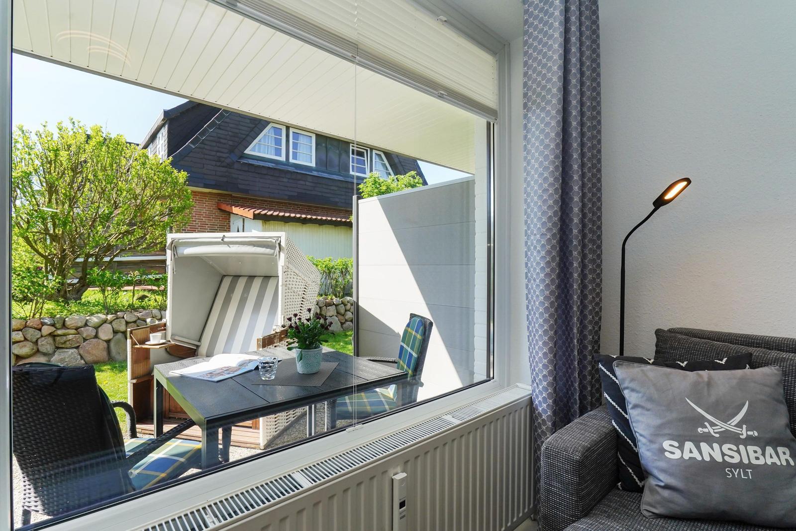 Durch großes Fenster Blick auf Garten mit Liegestuhl und Tisch. Im Raum Sofa mit Kissen und Stehlampe.