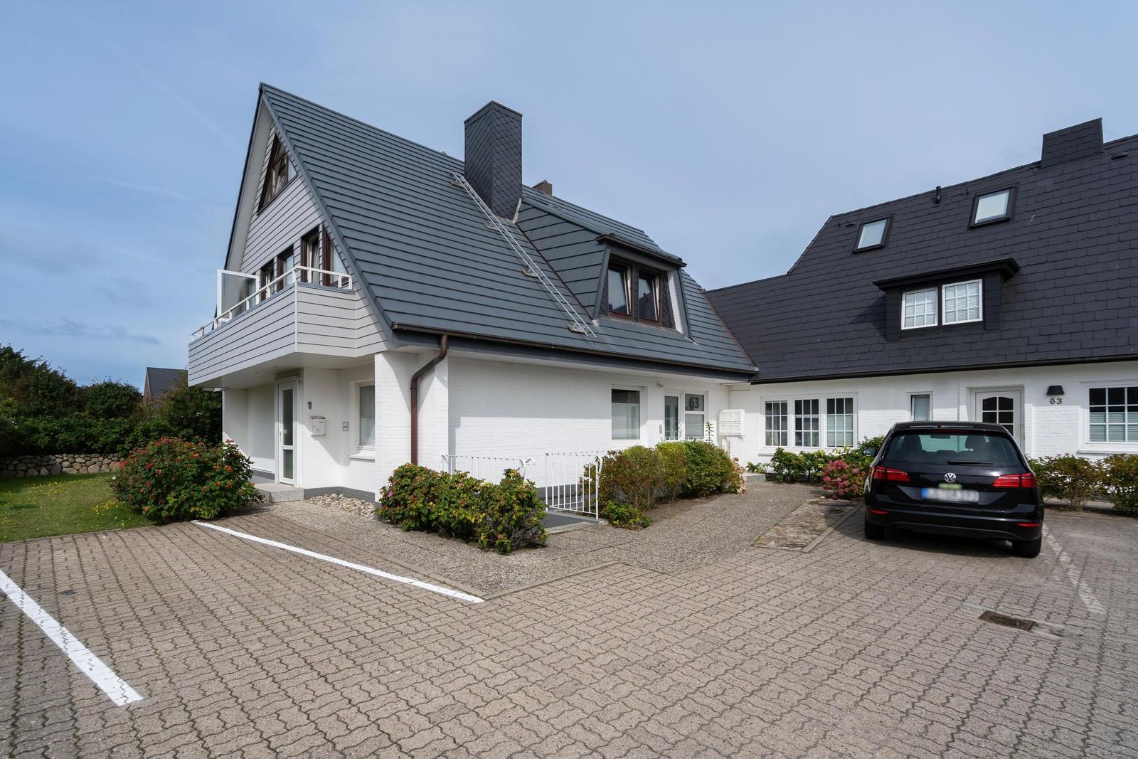 Ein weißes Haus mit dunklem Dach und Balkon. Vor dem Haus steht ein Auto.