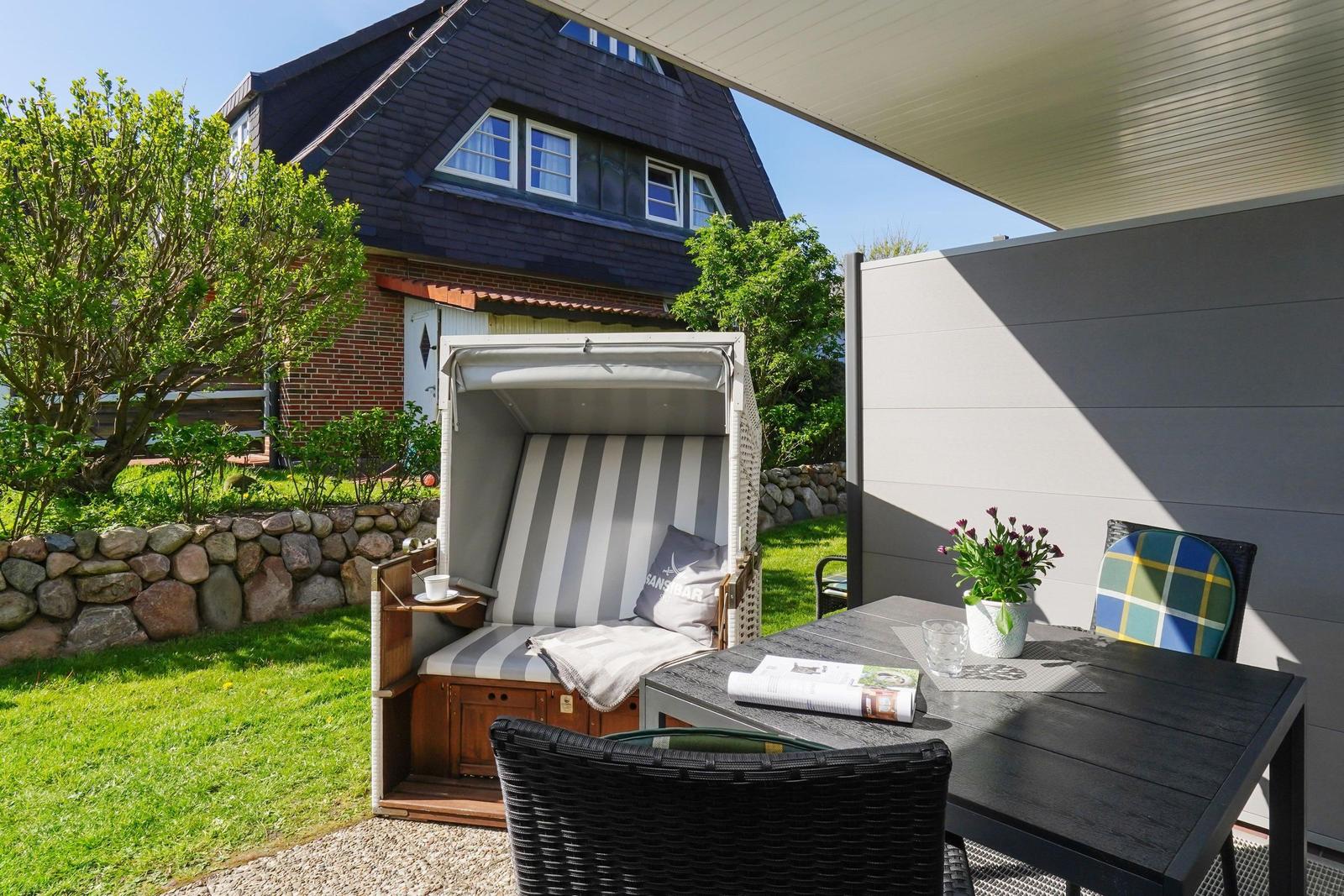 Terrasse mit Liege, Tisch und Stühlen. Hintergrund: Haus mit Steinmauer und Grün.