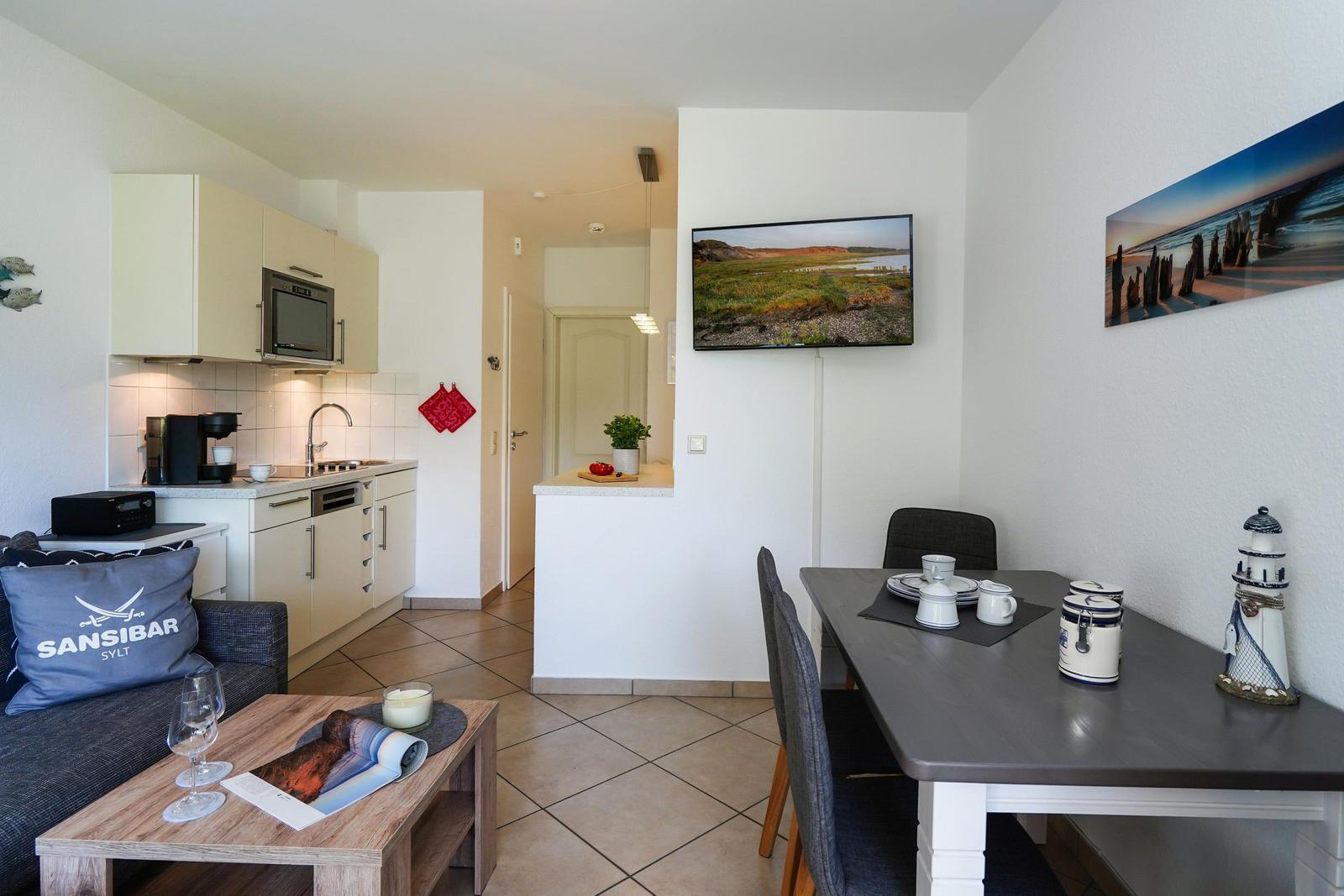 Wohnküche mit Esstisch, TV und Küchenzeile. Dekoration mit Leuchtturm und Landschaftsbild.