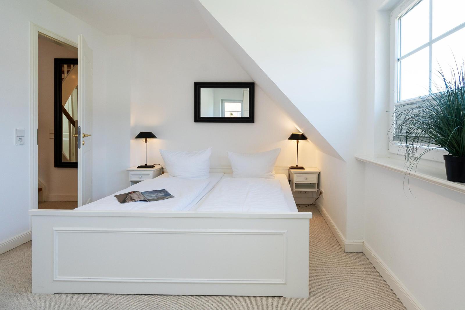 Schlafzimmer mit weißem Bett, zwei Nachttischen und Spiegel an schräger Wand.