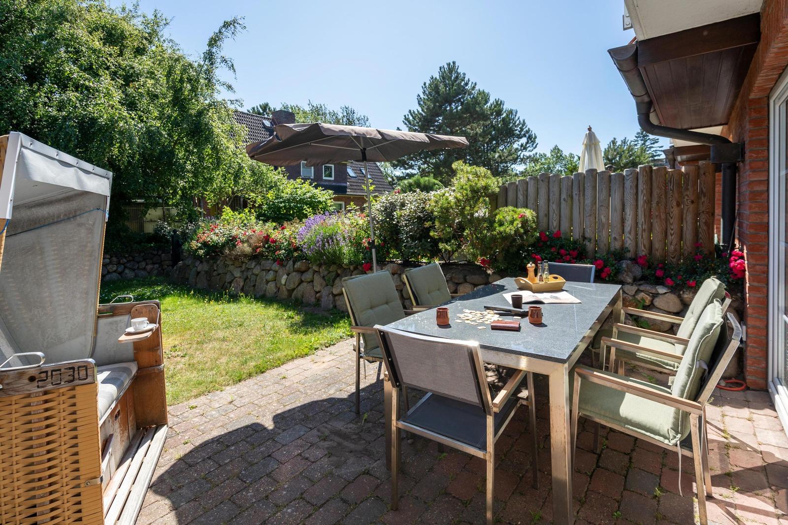 Terrasse mit Tisch, Stühlen und Sonnenschirm. Garten mit Blumen und Holzzaun.