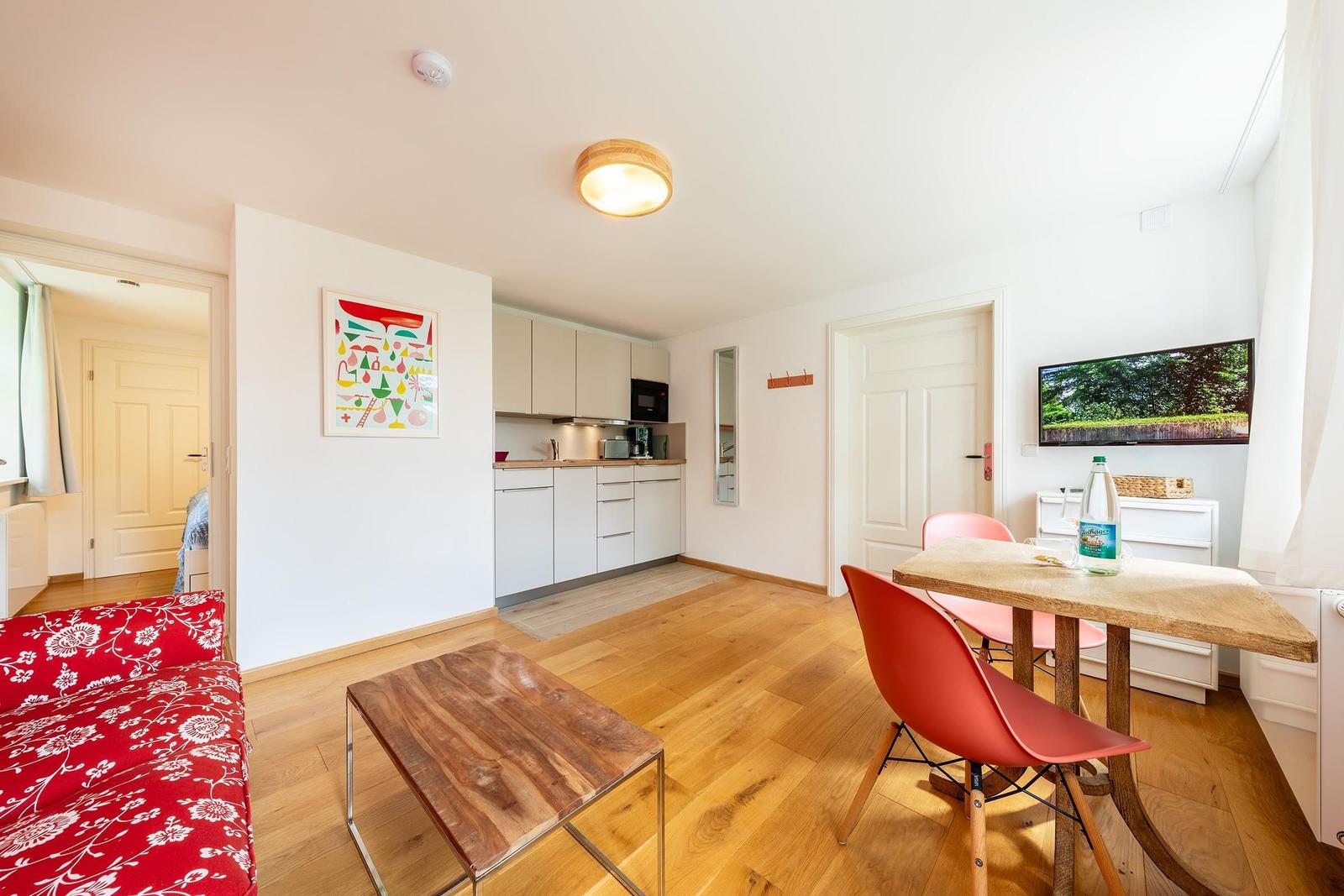 Wohnzimmer mit Essbereich, Küche und Fernseher. Holzboden und rote Sitzgelegenheiten.