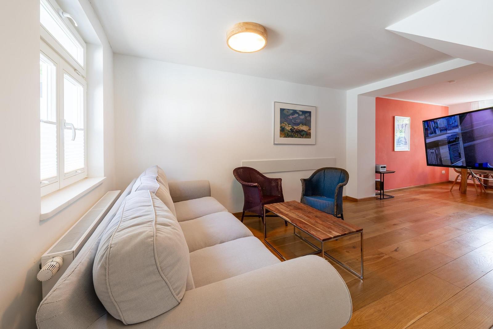 Wohnbereich mit Sofa, zwei Sesseln und Holztisch. An der Wand hängt ein Bild. Im Hintergrund ist ein Fernseher.