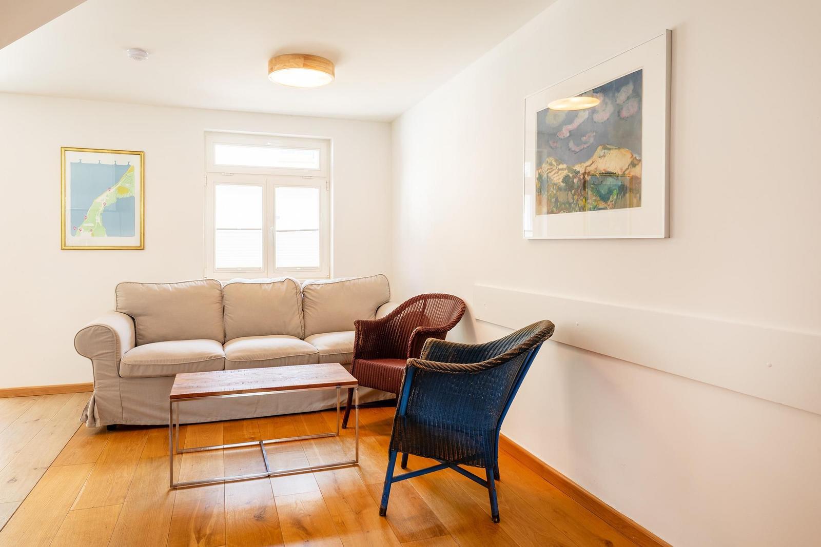 Wohnzimmer mit Sofa, zwei Stühlen und Holzboden. An den Wänden hängen Bilder.