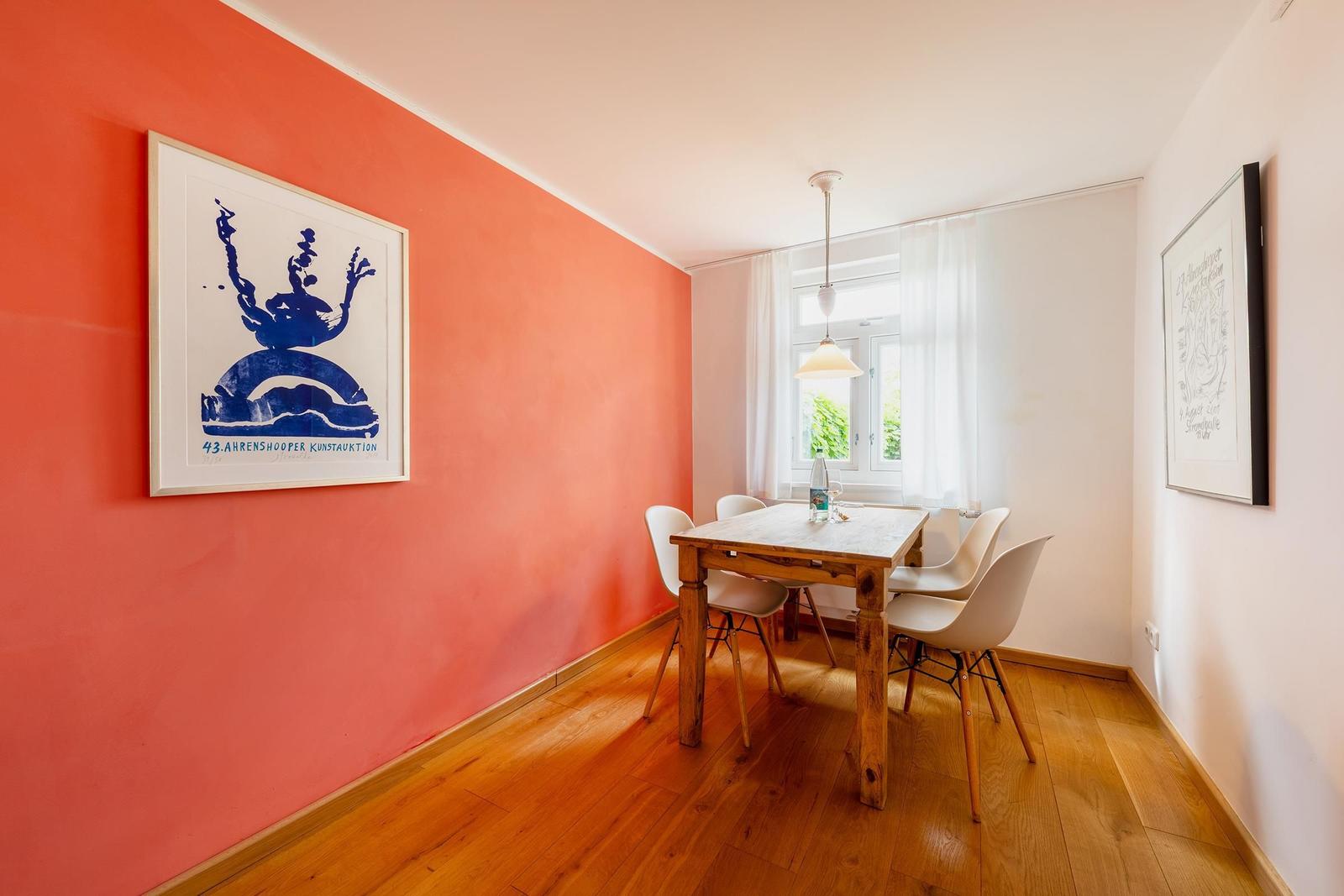 Esszimmer mit Holztisch, weißen Stühlen und orangefarbener Wand. Fenster mit weißen Vorhängen.