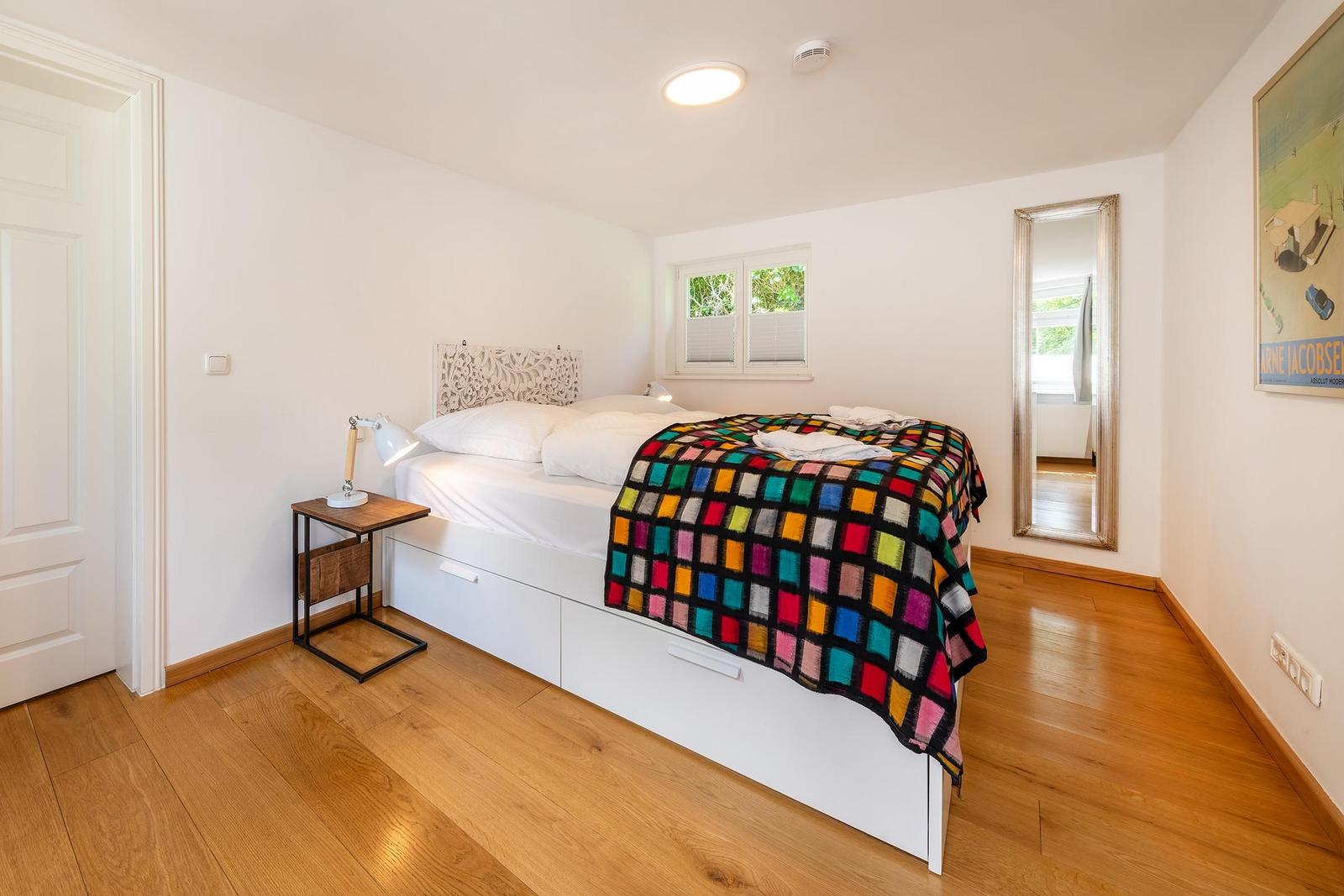 Schlafzimmer mit Bett, Nachttisch und Spiegel. Holzfußboden und Fenster mit Blick.