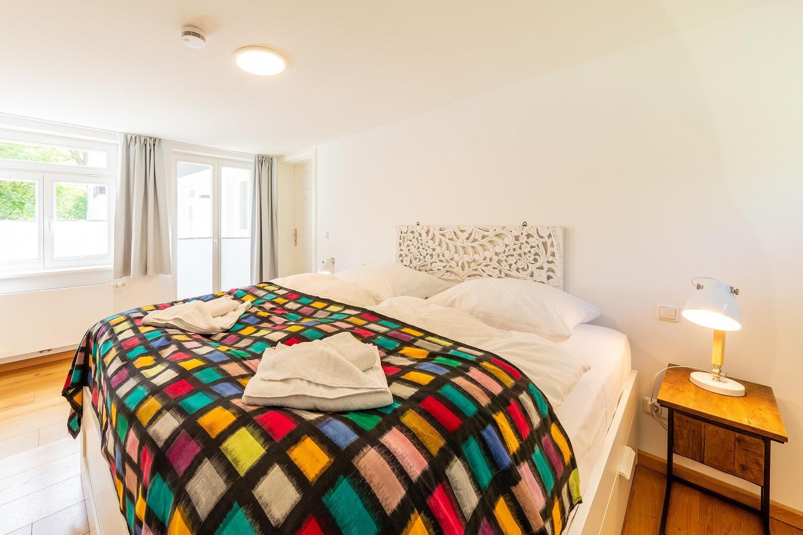Schlafzimmer mit Bett, buntem Deckenbezug und Nachttisch mit Lampe.