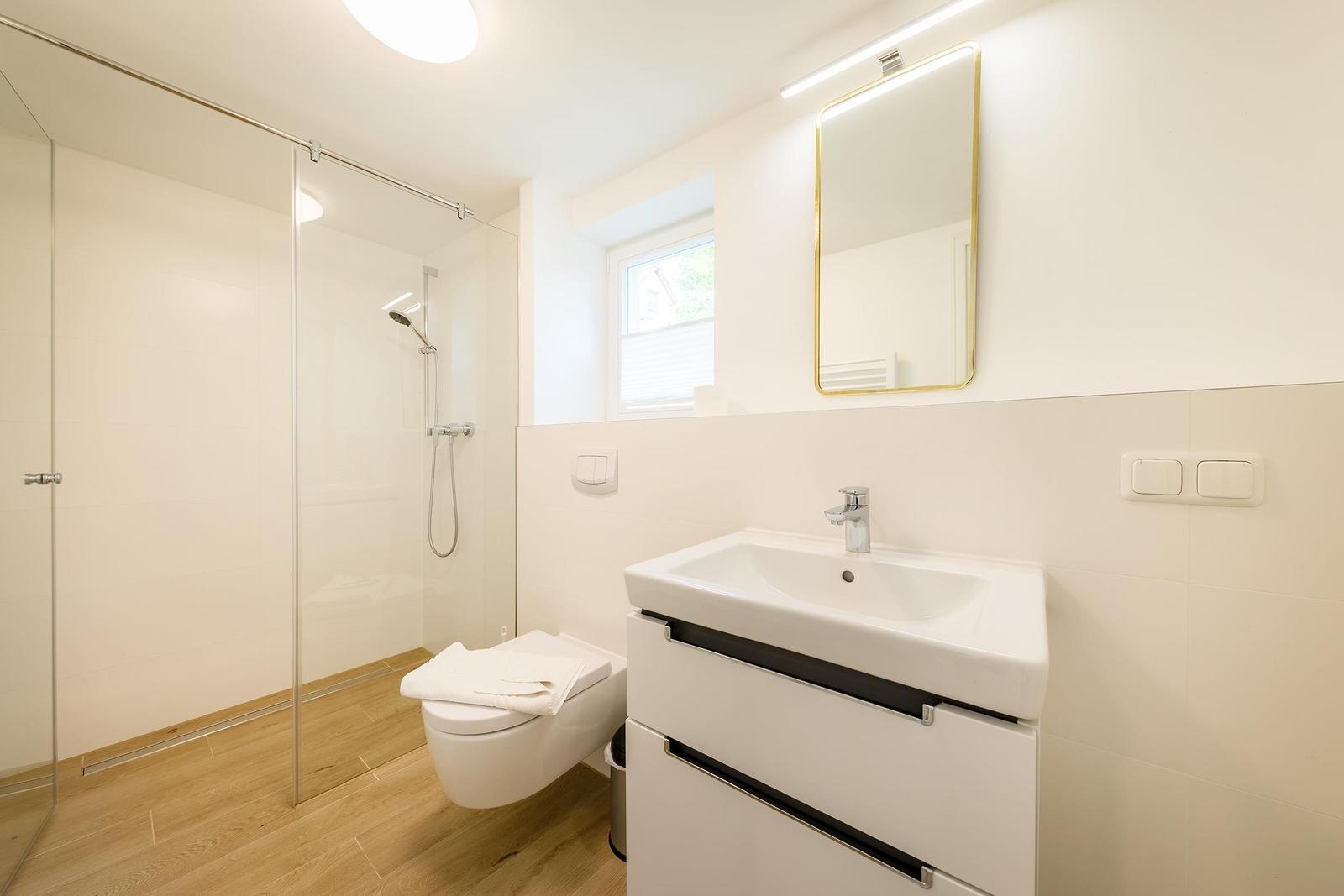 Badezimmer mit Dusche, Waschbecken und Toilette. Holzfußboden und Fenster.
