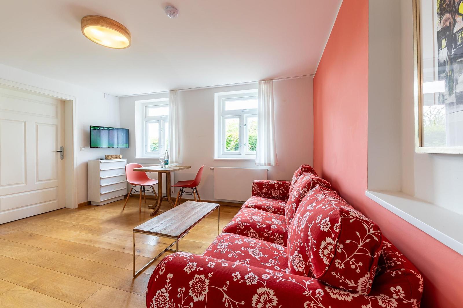 Ein Wohnzimmer mit rotem Sofa, Holzboden und zwei Fenstern.