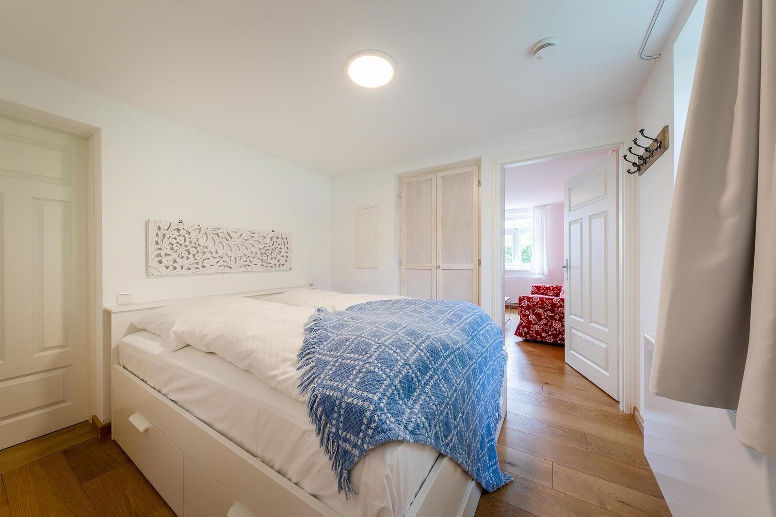 Schlafzimmer mit Bett, blauer Decke und Holzboden. Tür führt in Wohnbereich mit rotem Sessel.
