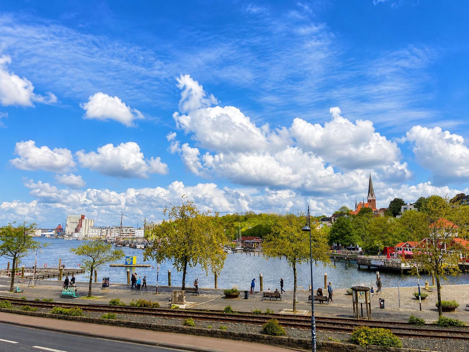 Hafenstadt mit Blick auf Wasser, Bäume und Kirche unter blauem Himmel mit weißen Wolken.