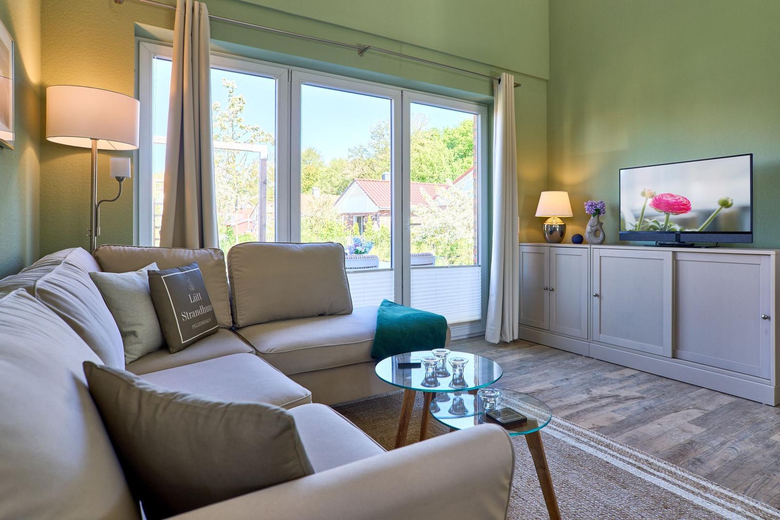 Wohnzimmer mit Sofa, Glas-Tisch und TV. Große Fenster mit Blick auf Garten.