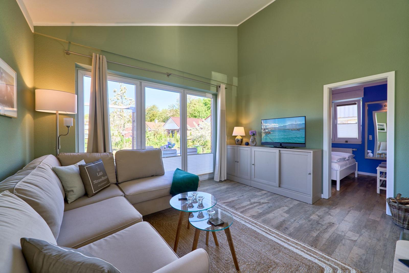 Wohnzimmer mit Sofa, Glas-Tisch und TV. Durchgang zu Schlafzimmer mit blauer Wand.