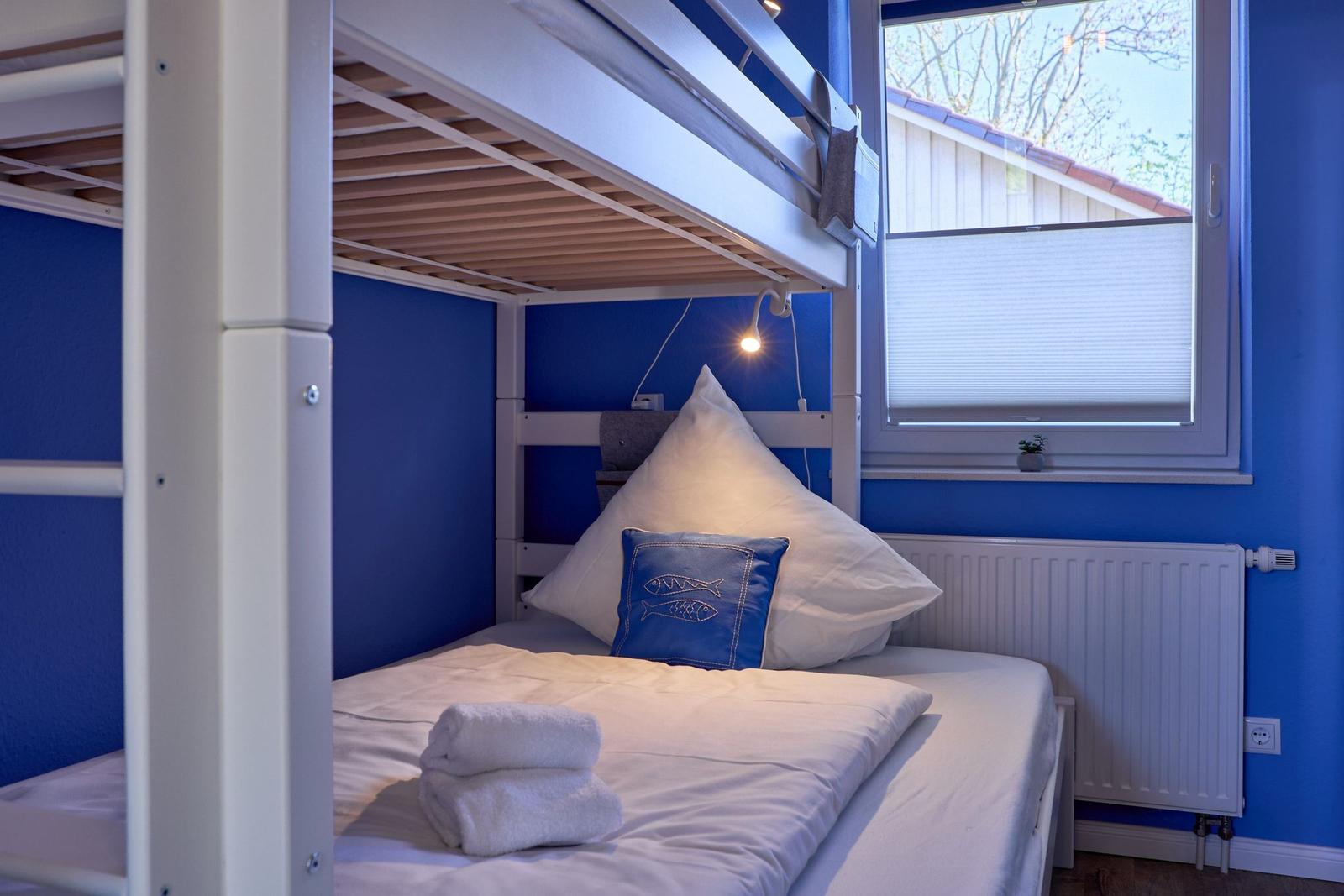 Ein Bett mit weißer Bettwäsche und blauem Kissen unter einem Doppelstockbett. An der Wand hängt eine Lampe.