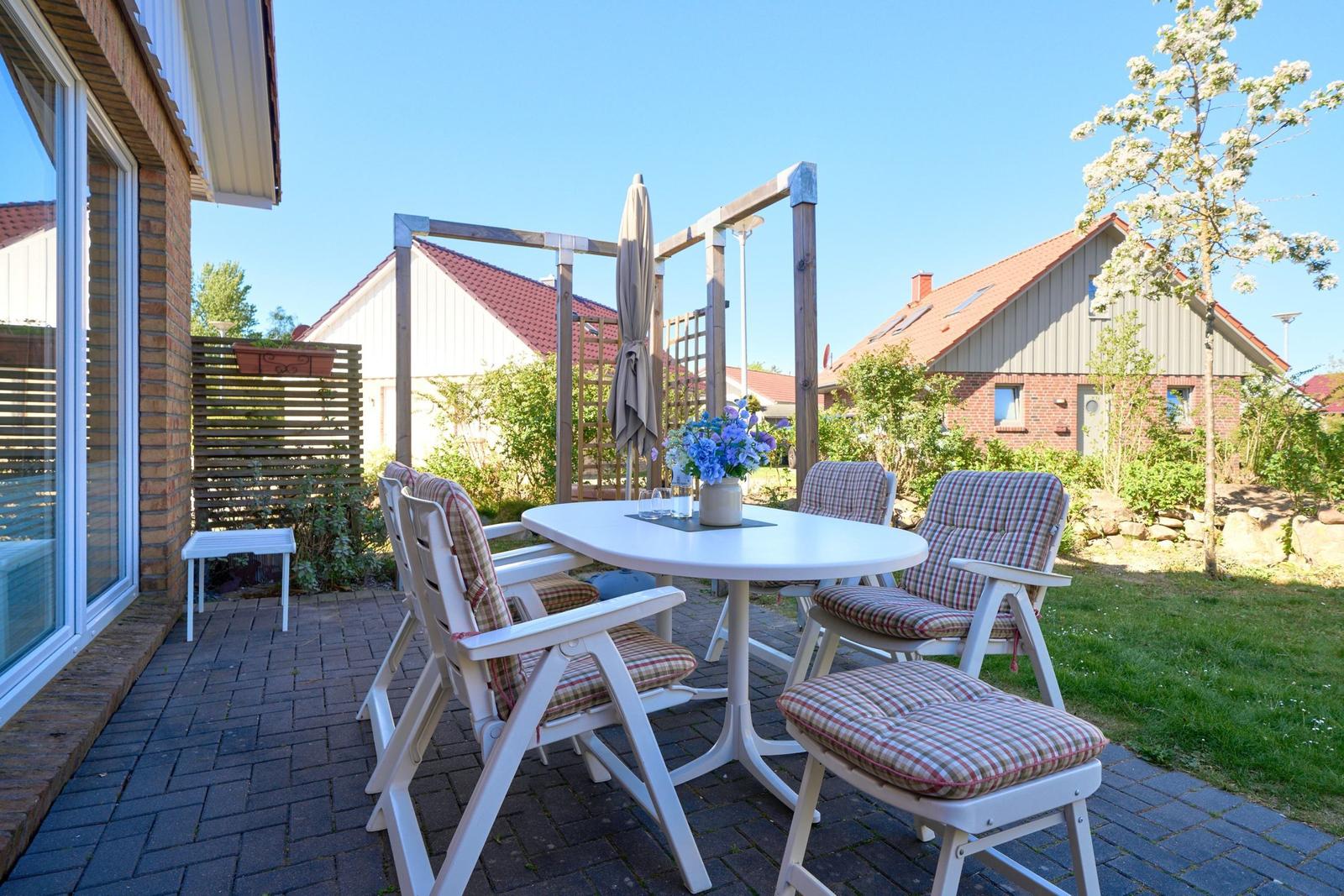 Terrasse mit weißem Tisch, Stühlen und Sonnenschirm. Hintergrund: Garten und Nachbarhäuser.
