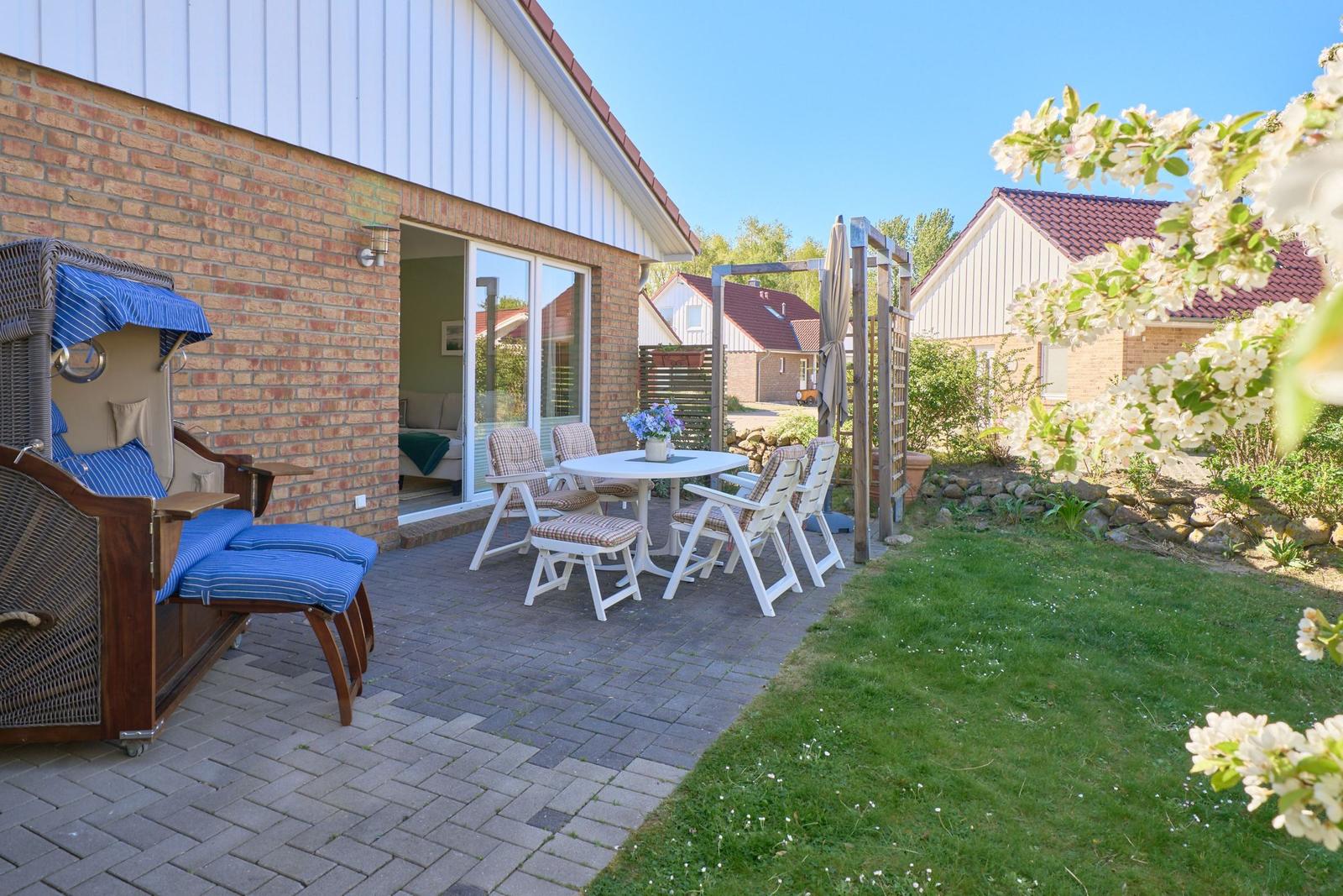 Terrasse mit Gartenhaus, Tisch, Stühlen und Liegestuhl. Blick auf grünen Rasen und blühende Sträucher.