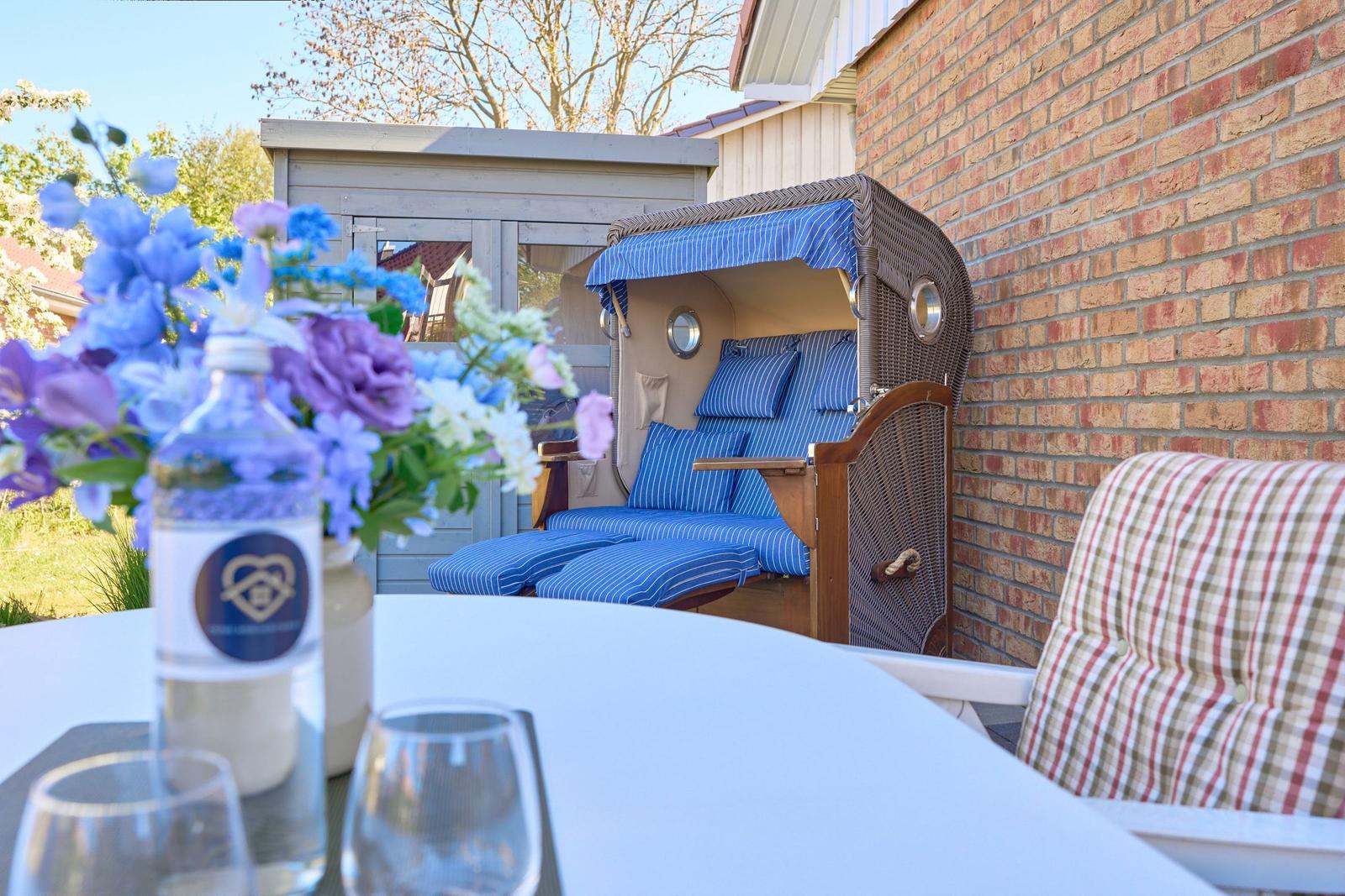 Terrasse mit Strandkorb, Tisch, Stühlen und Blumenarrangement.