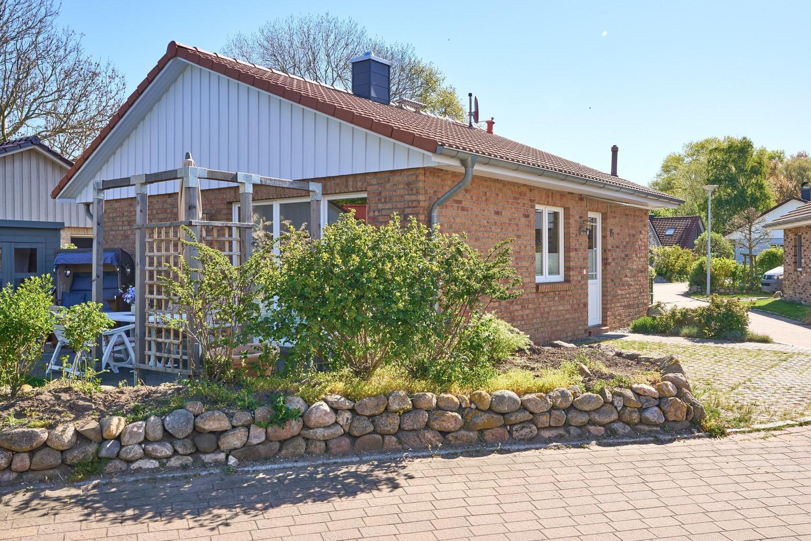 Ein kleines Haus mit Steinmauer, Garten und Terrasse unter blauem Himmel.