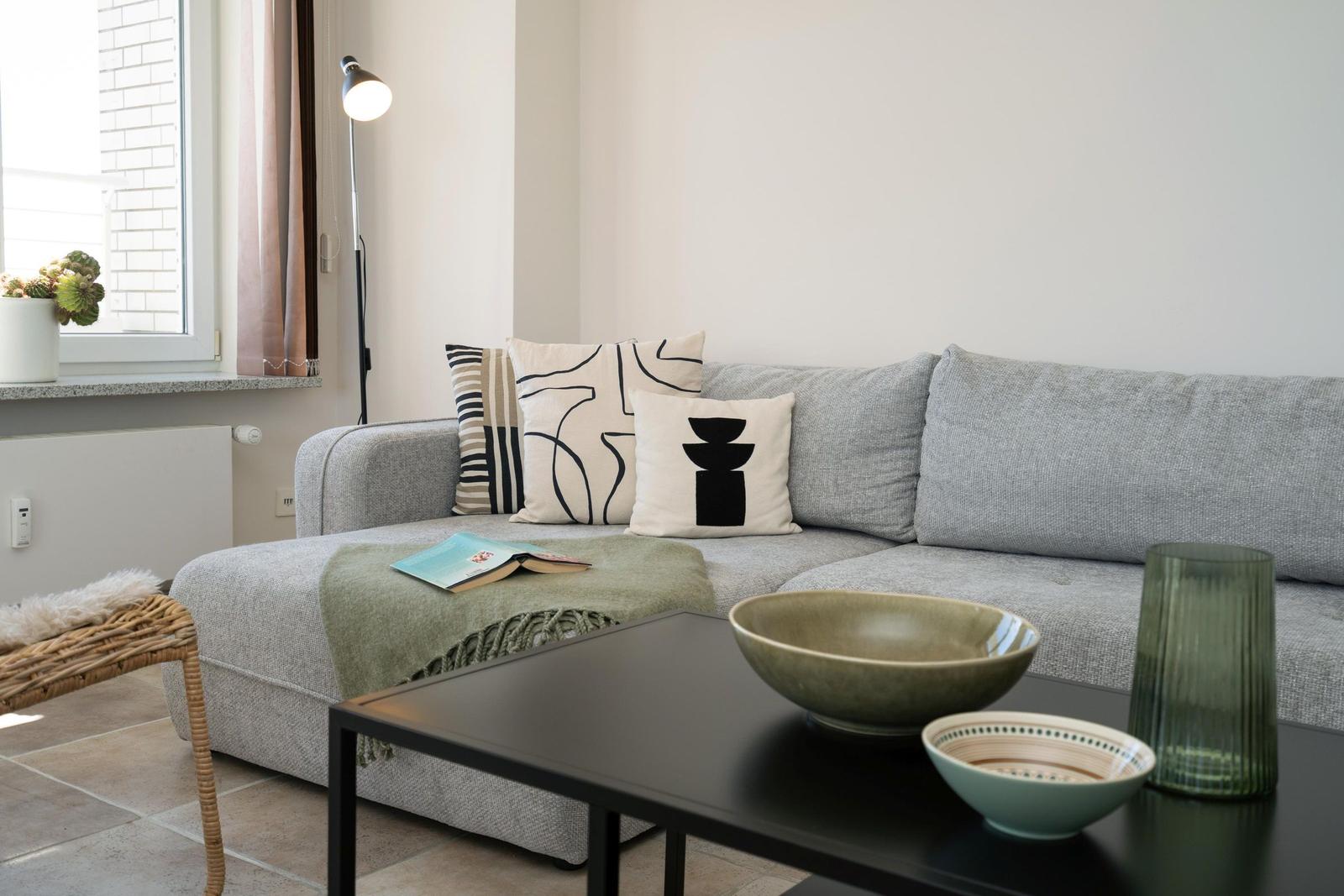 Ein grauer Sofa mit Kissen und Tisch im Vordergrund. Auf dem Tisch stehen Schalen und ein Glas.