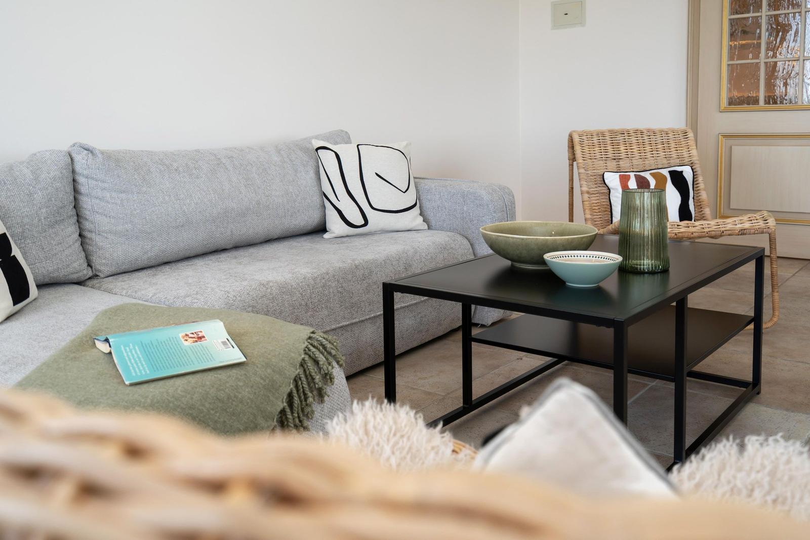 Ein grauer Sofa mit Kissen, ein schwarzer Tisch und eine Rattan-Stuhl.