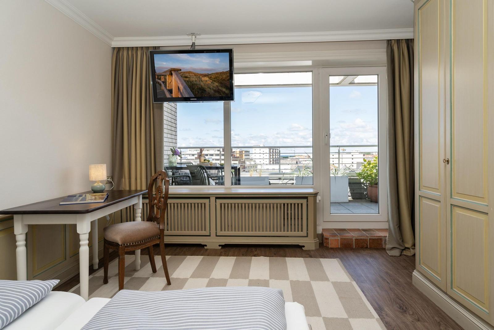 Zimmer mit Schreibtisch, Fernseher und Balkonblick auf die Stadt.