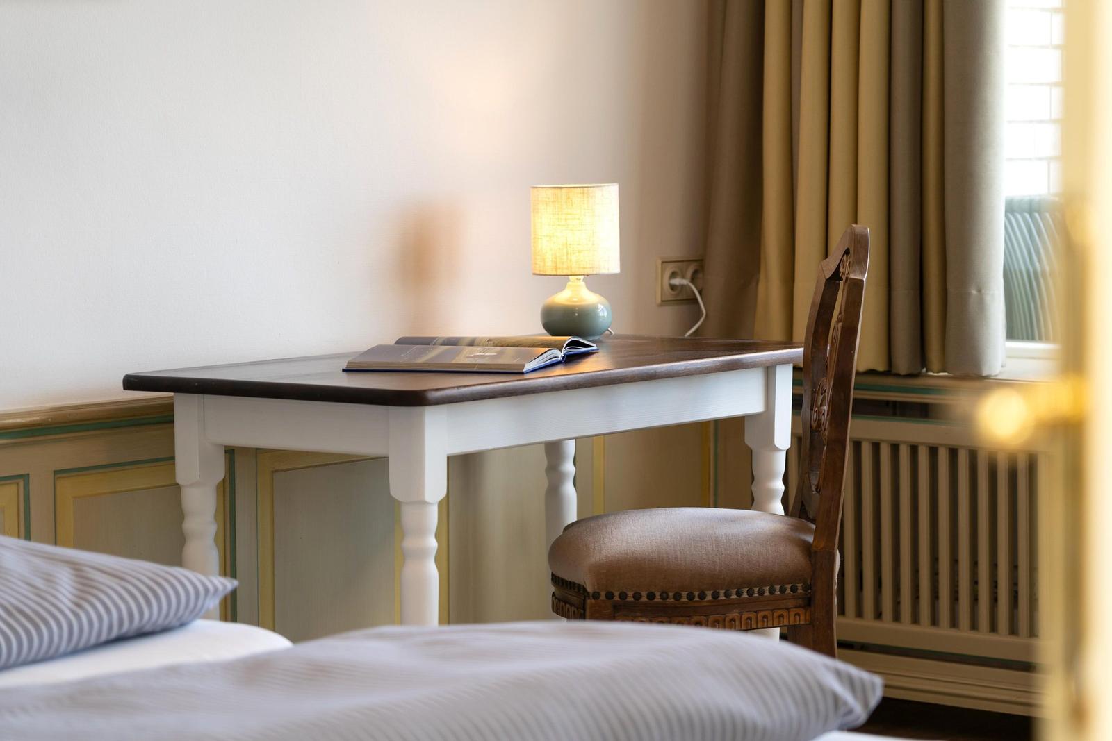 Schreibtisch mit Stuhl, Lampe und Buch neben Fenster mit Vorhängen.
