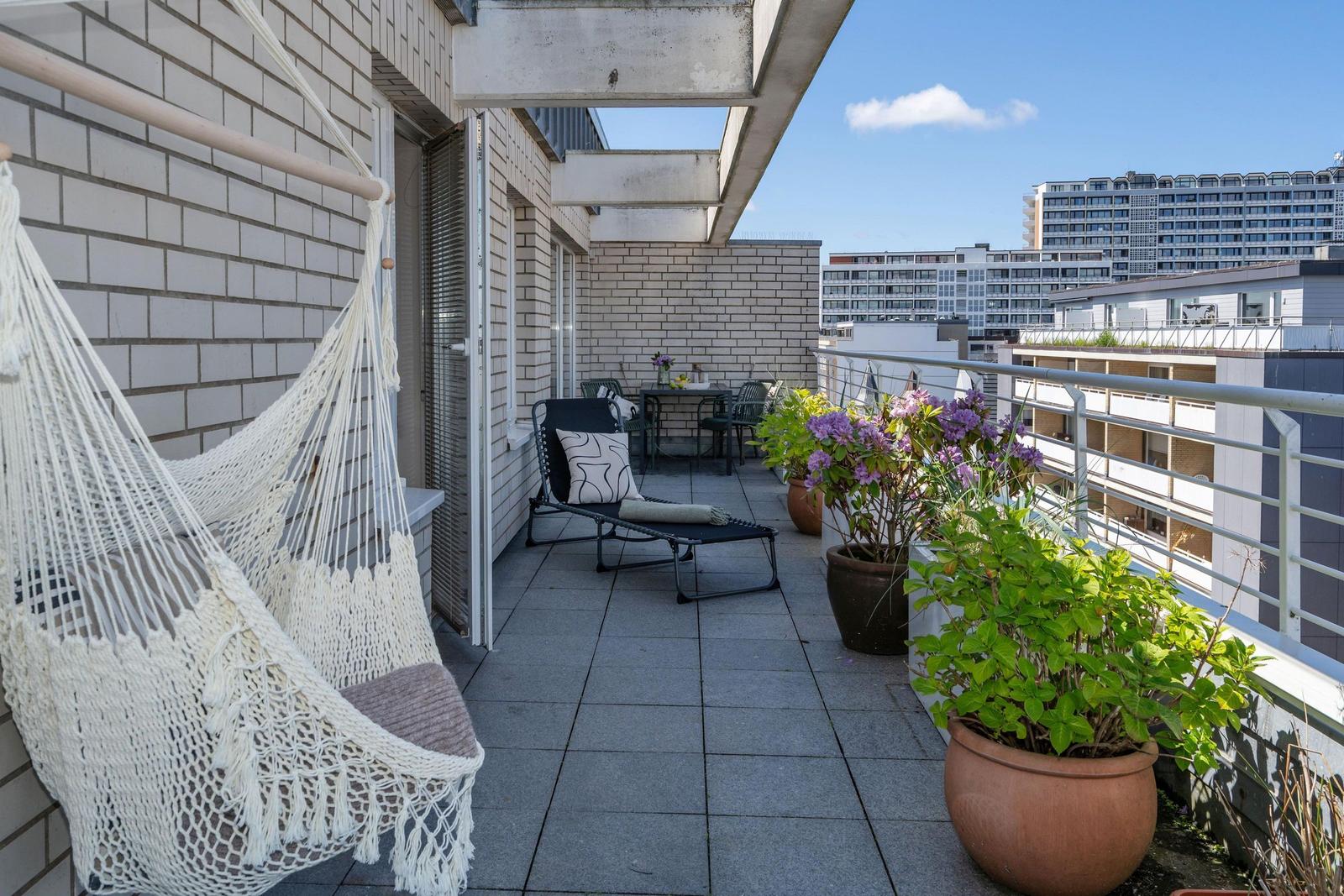Terrasse mit Hängematte, Liegestuhl und Pflanzen. Blick auf Hochhäuser.