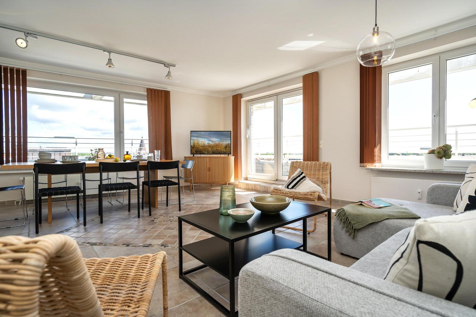 Wohnbereich mit Sofa, Esstisch und TV. Große Fenster und Balkon.