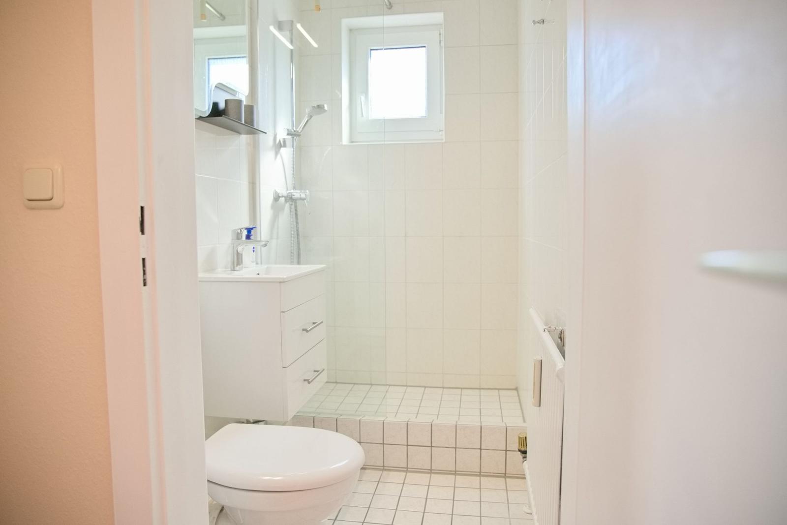 Badezimmer mit Dusche, Waschbecken und Toilette. Weiße Fliesen und Fenster.