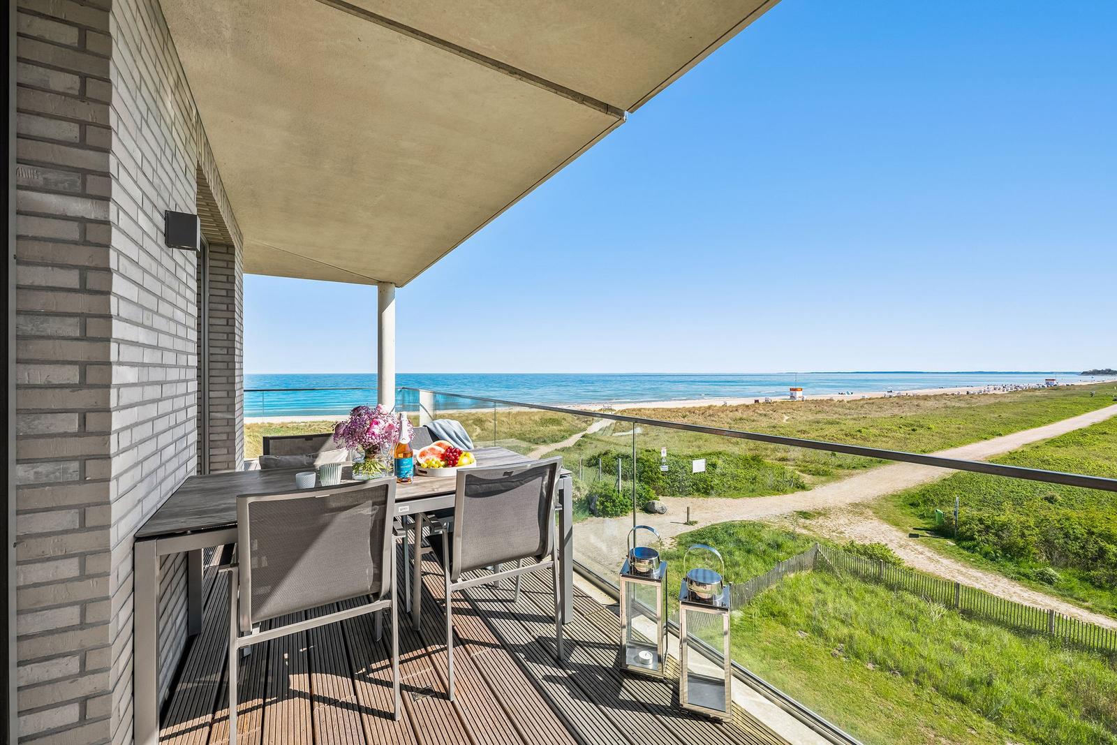 Terrasse mit Tisch, Stühlen und Meerblick. Glasgeländer und Holzdielen.