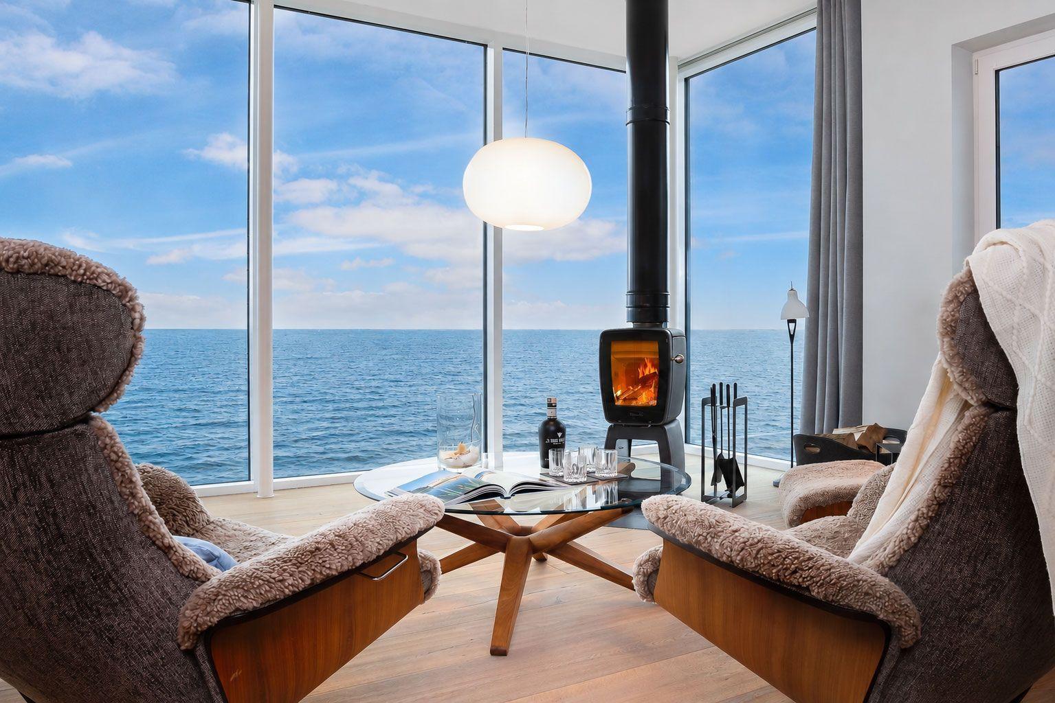 Zwei Sessel um einen Tisch mit Kamin und Meerblick durch große Fenster.