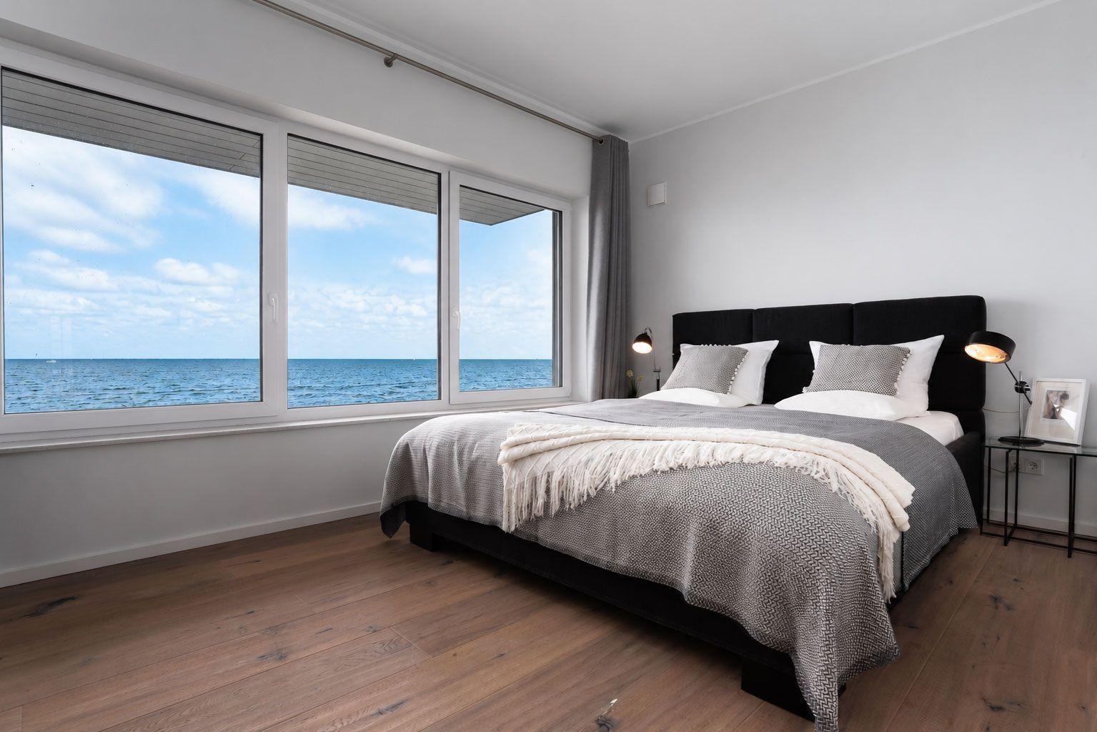 Schlafzimmer mit Blick auf das Meer durch große Fenster. Schwarzes Bett, Holzboden, Nachttische.